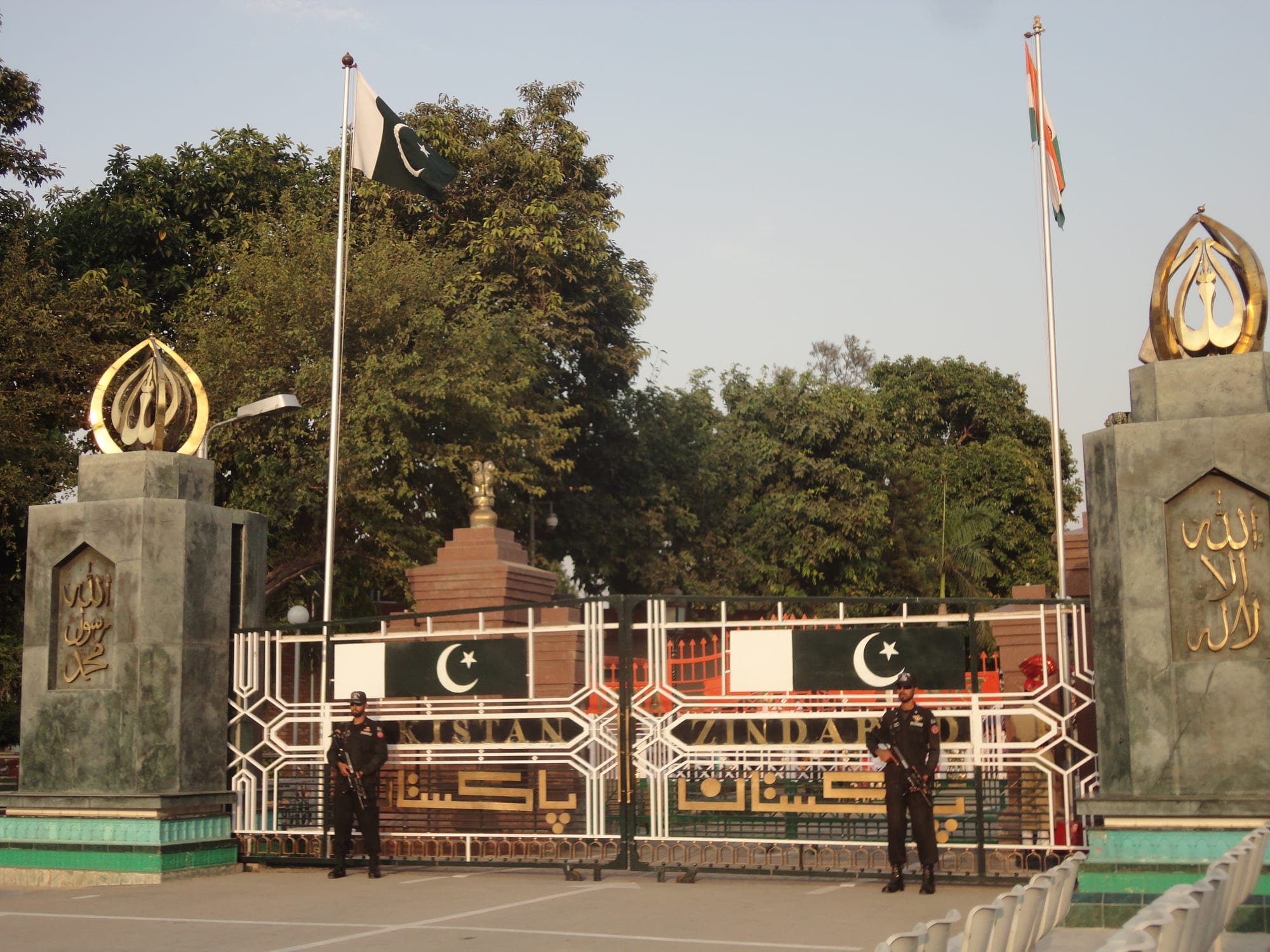 Two armed guards in black uniforms standing before the closed white border gate at Wagah, with the Pakistani flag on one pole and the Indian flag on another