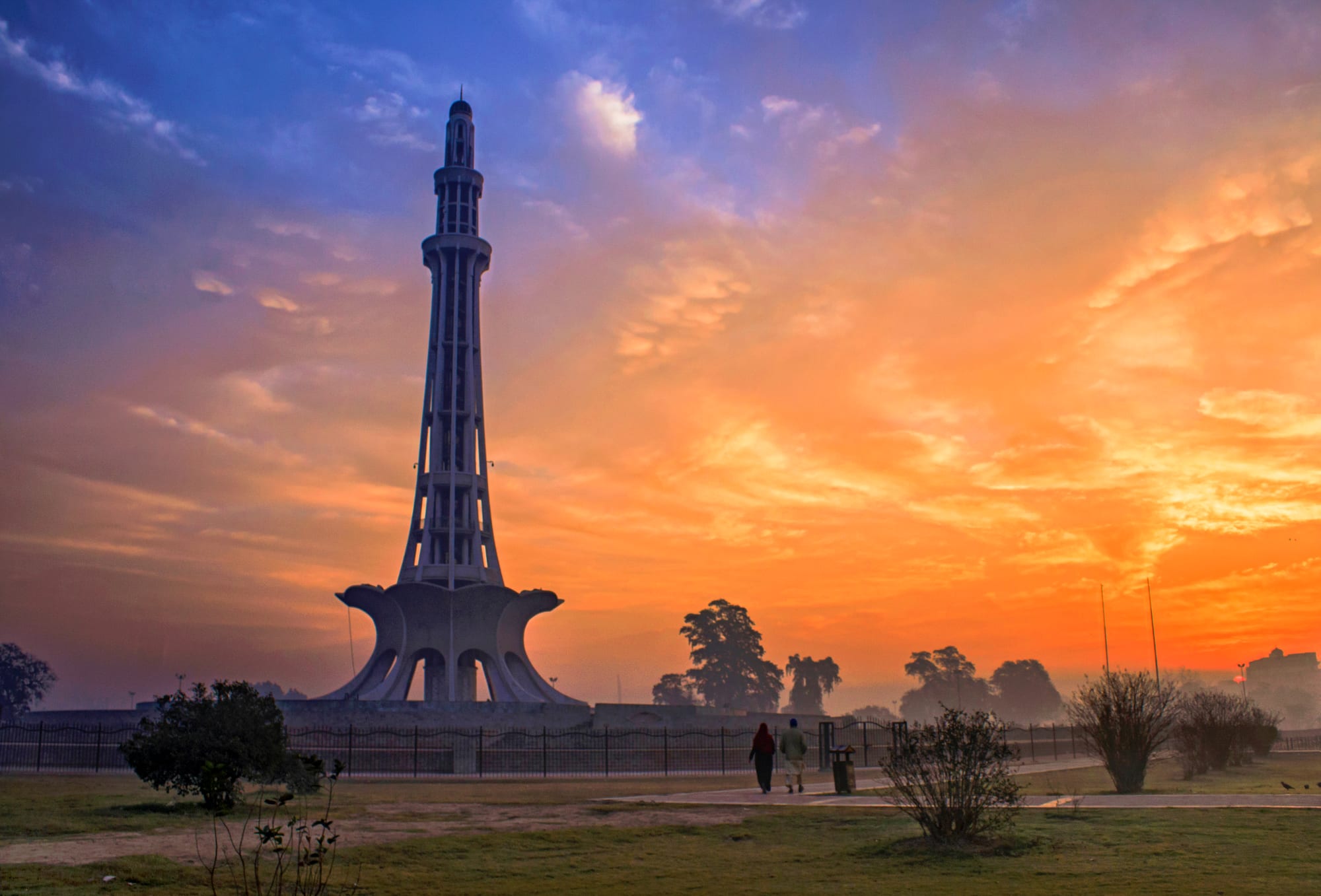 The tall white Minar-e-Pakistan tower rising from its flower-shaped base in a Lahore park, silhouetted against an orange and blue sky