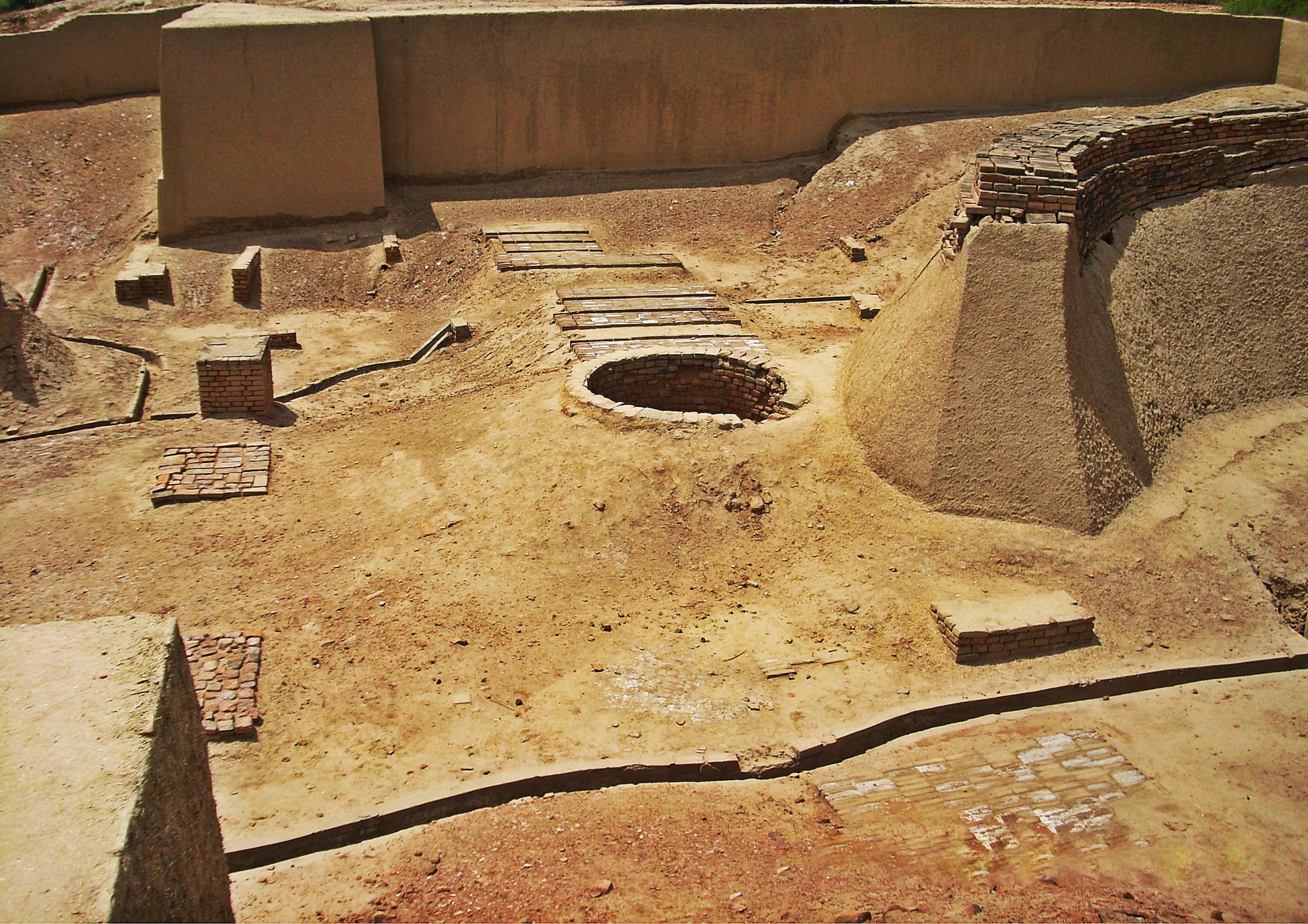 Excavated ruins at Harappa with a round brick-lined well, low brick platforms and narrow channels set in pale earth
