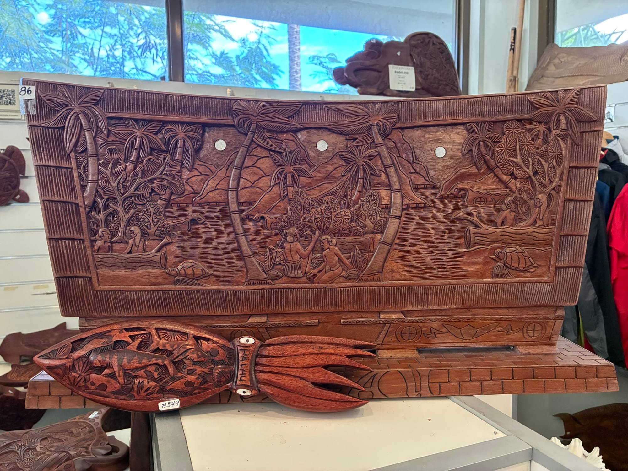 A large dark wooden storyboard deeply carved with palm trees, people, turtles and a dolphin by the sea, displayed on a stand with a smaller carved fish-shaped board in front of it