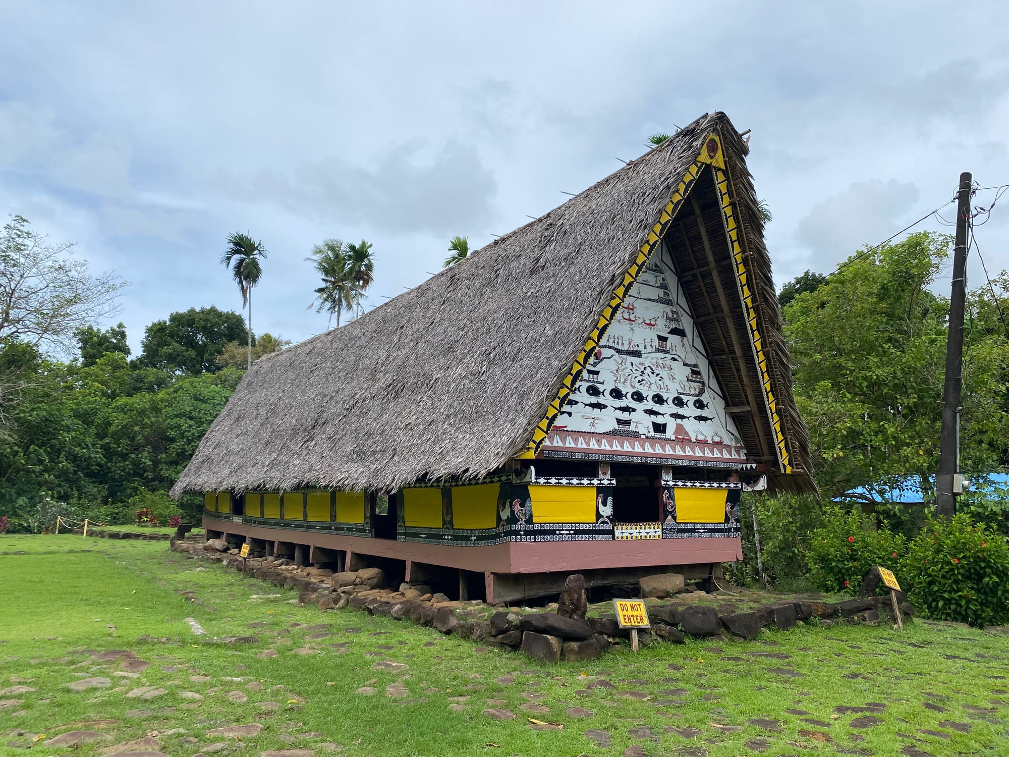 A long traditional meeting house with a steep, high-peaked thatched roof, a gable painted with rows of figures, fish and symbols, and yellow walls decorated with rooster motifs, standing on a stone platform on a lawn