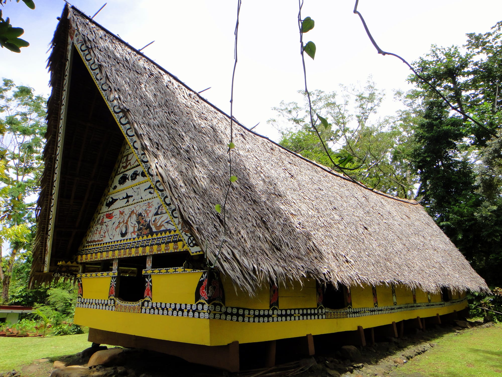 A traditional Palauan bai with a steeply pitched thatched roof and a brightly painted yellow gable showing legends and figures