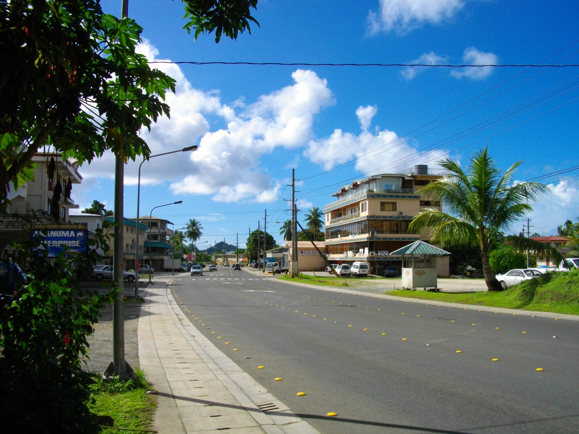 A wide street in downtown Koror lined with low buildings, palm trees and parked cars under a bright blue sky