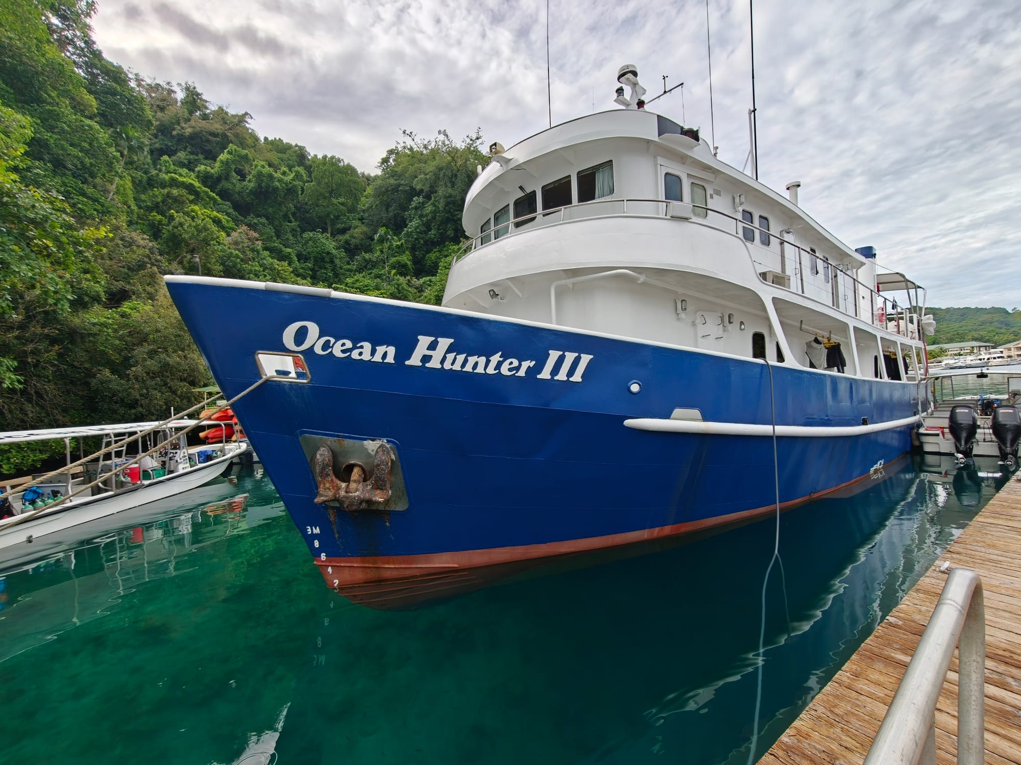 A liveaboard dive boat named Ocean Hunter III docked at a jetty in Koror, with smaller dive boats alongside and forested hills behind