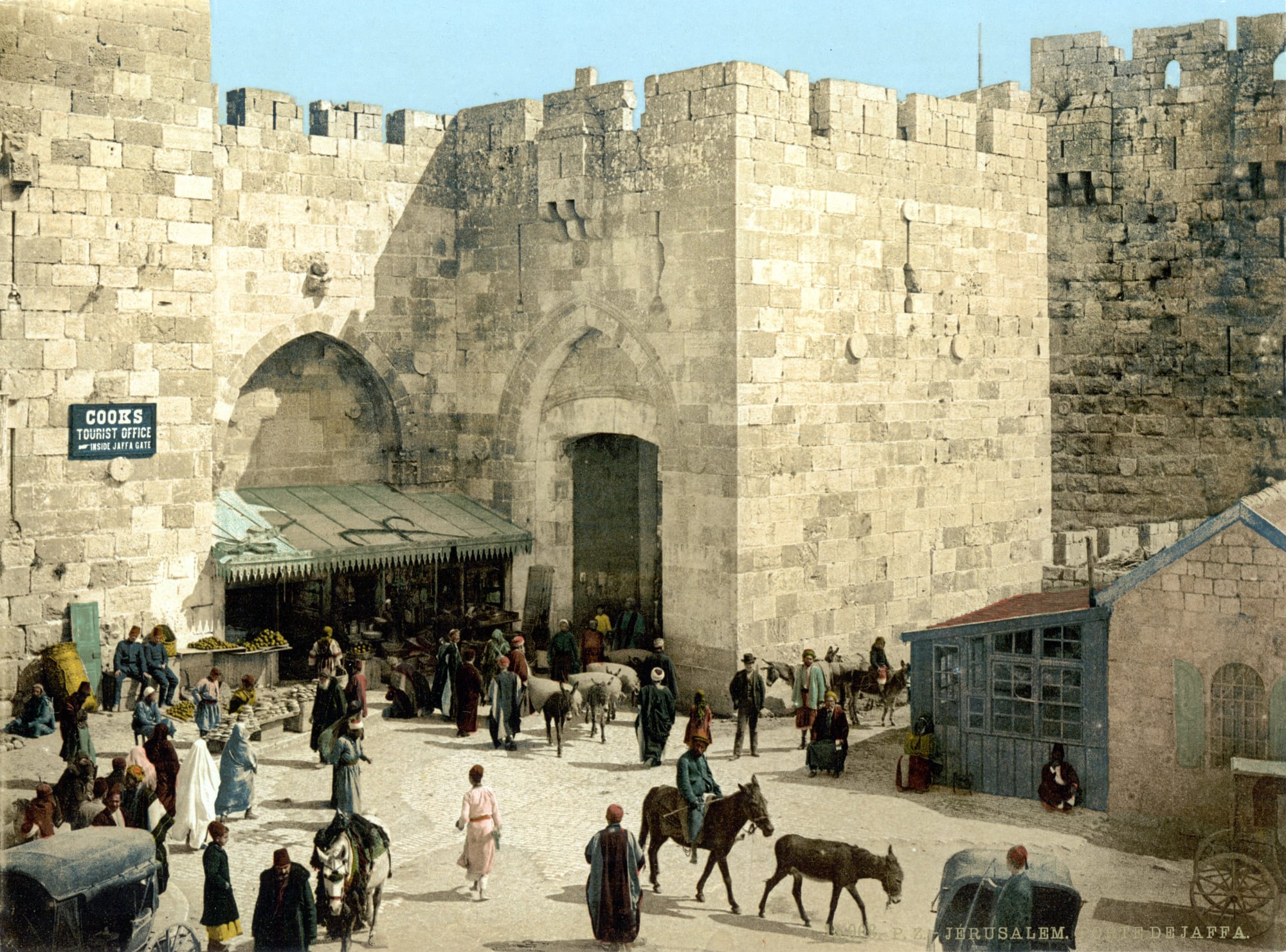 An early colour photograph of the stone Jaffa Gate in Jerusalem's city wall, with market stalls, donkeys and people in robes and fezzes on the open ground in front