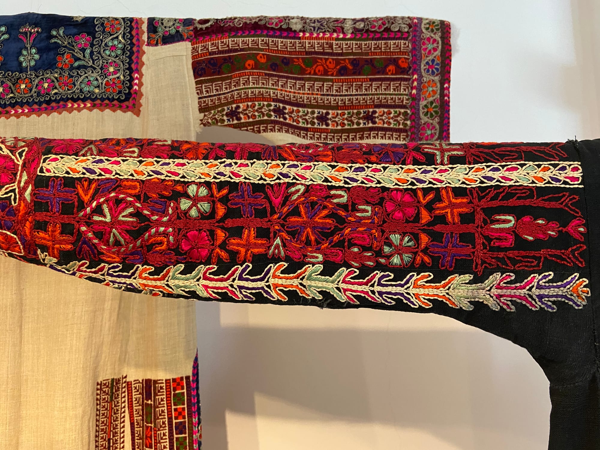 Two embroidered dress sleeves on display, the front one of black cloth covered in red, orange, pink and pale green cross-stitch flowers and borders, the one behind on pale linen with bands of geometric and floral patterns