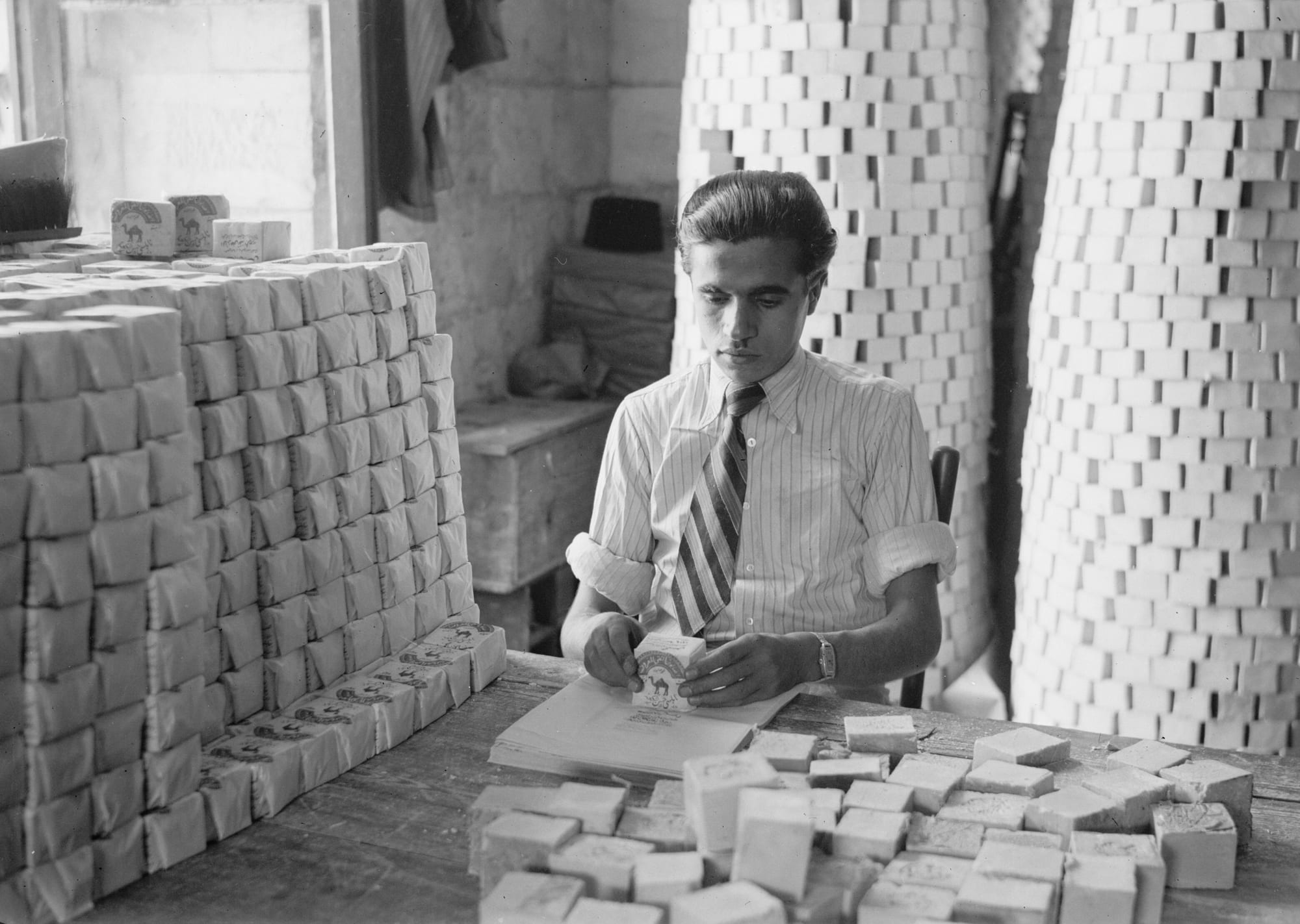 Black and white photograph of a young man in a shirt and tie seated at a table, wrapping bars of soap in printed paper, with tall cylindrical stacks of soap bars behind him