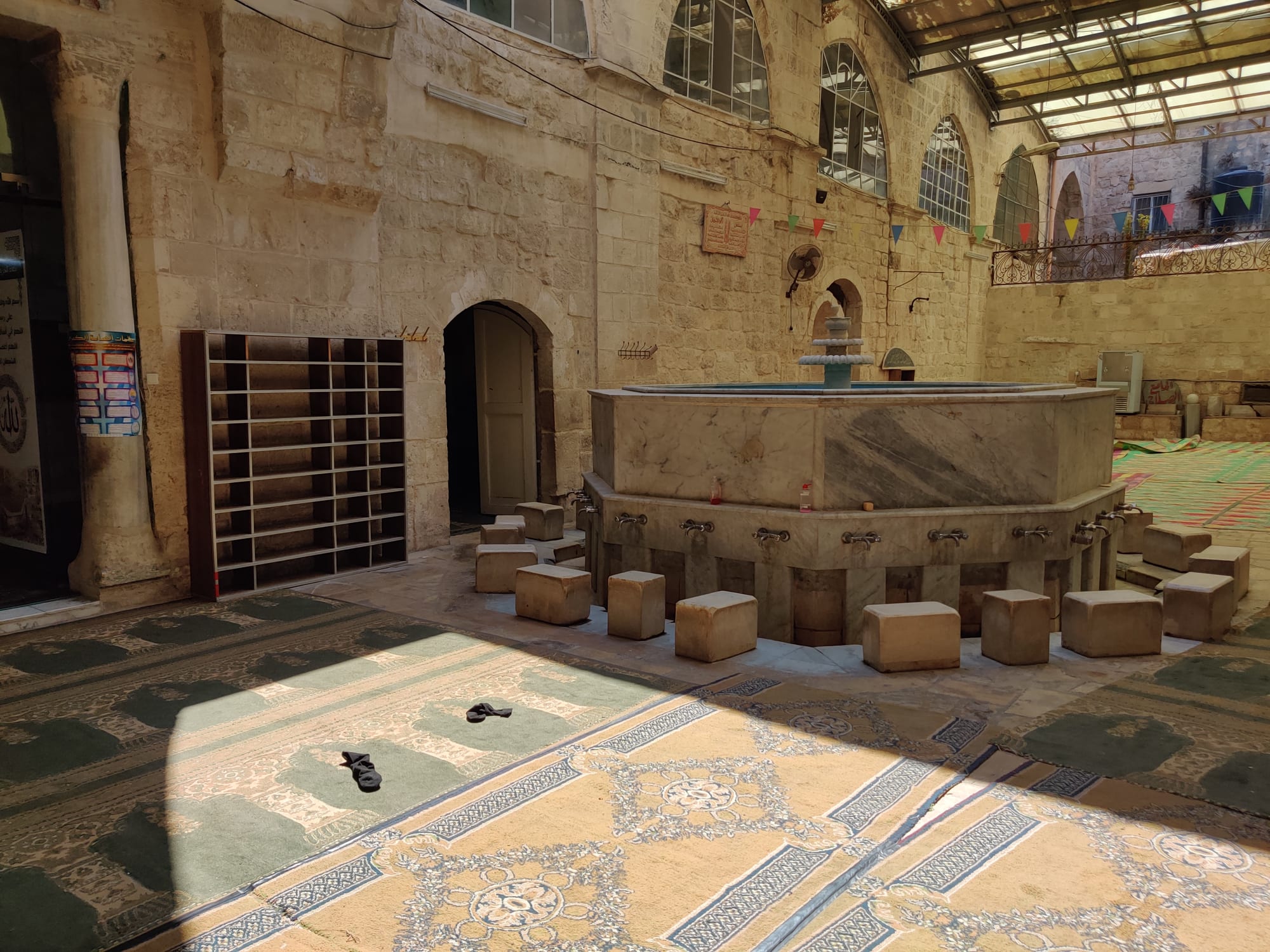 A stone mosque courtyard with an octagonal marble ablution fountain surrounded by low stone seats and taps, patterned carpets on the floor and shoe shelves beside an arched doorway