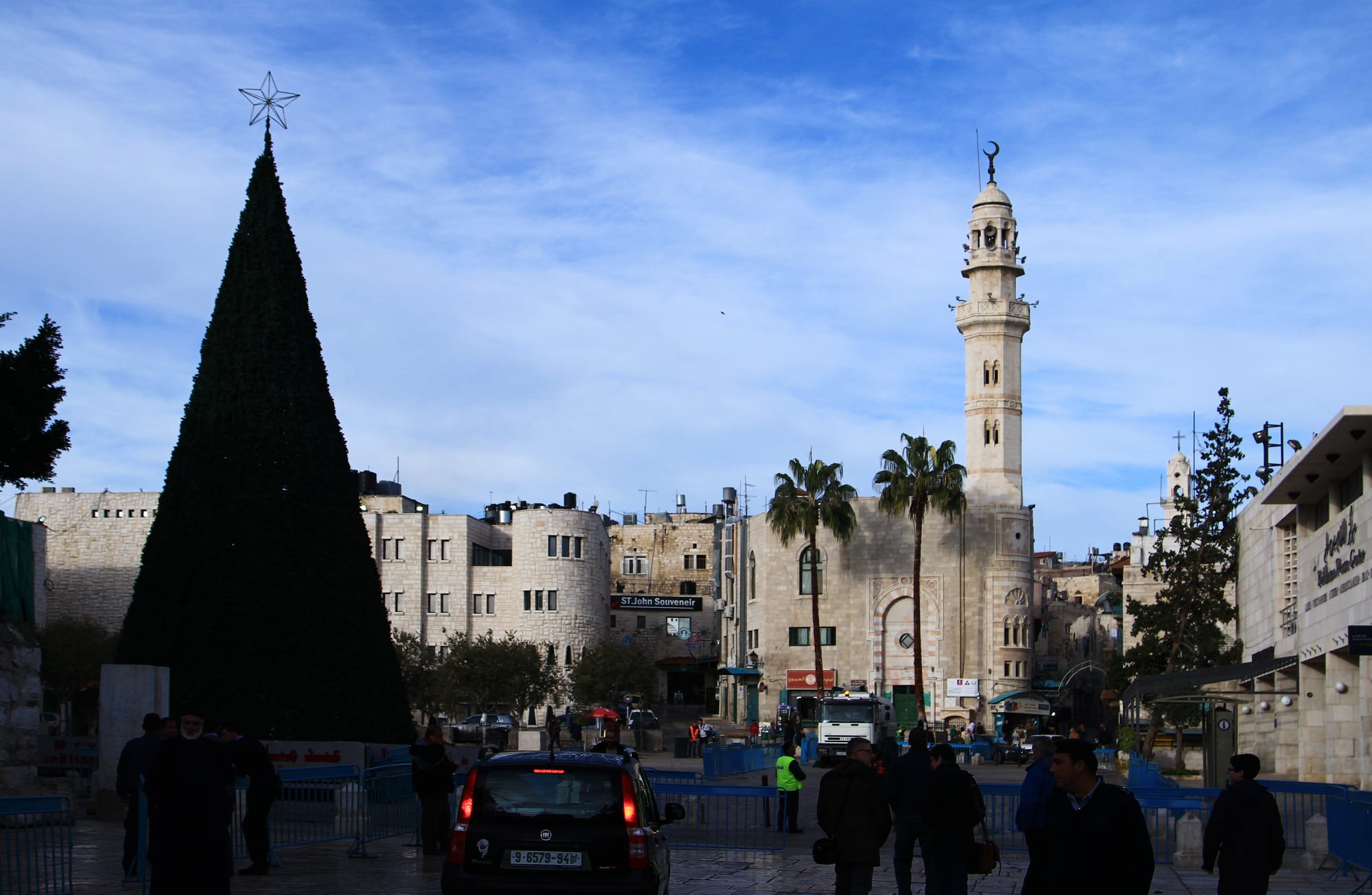 A tall dark conical Christmas tree topped with a star stands in a paved stone square, with stone buildings, palm trees and a tall mosque minaret beyond and a few people and cars in the foreground