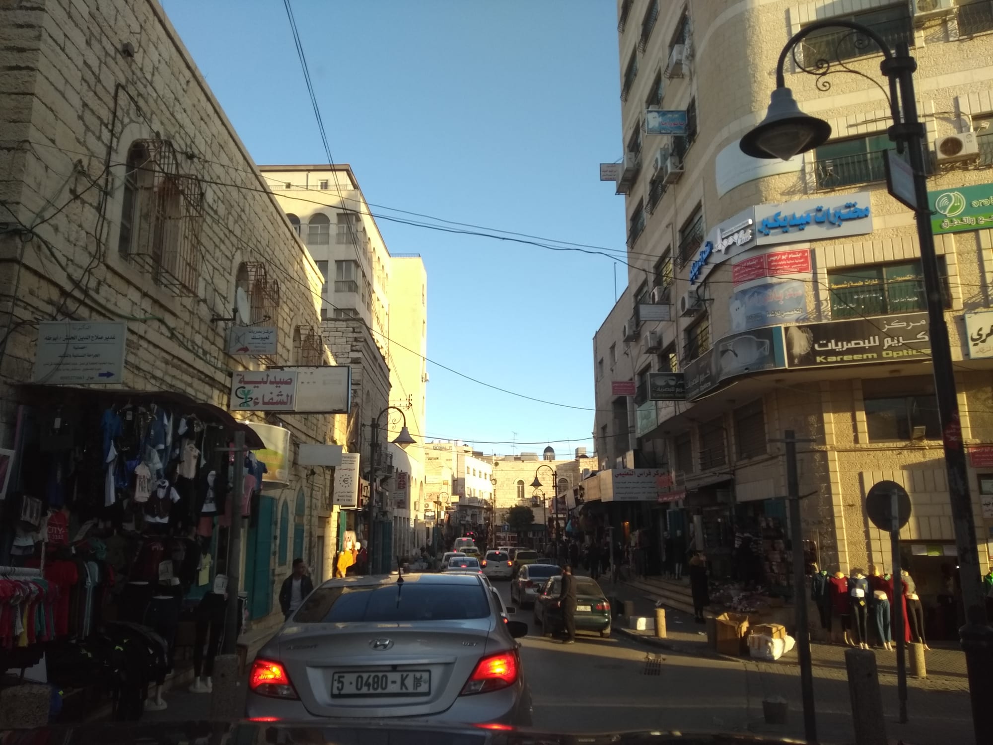 A narrow street lined with clothing shops and stone buildings in Bethlehem, cars queued along the road and shoppers on the pavement in late afternoon light