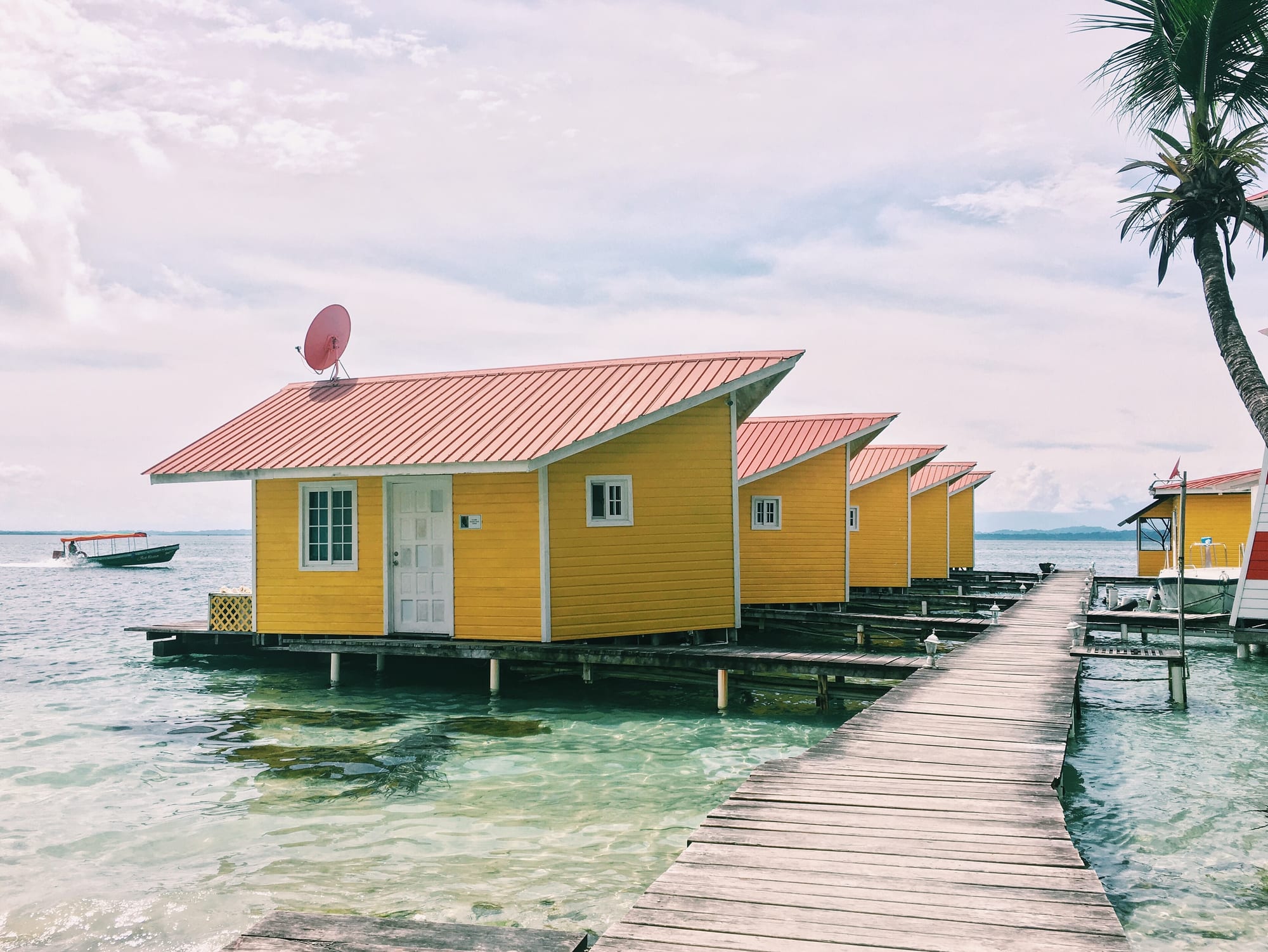 Colourful wooden cabins on stilts over turquoise water beside a palm tree