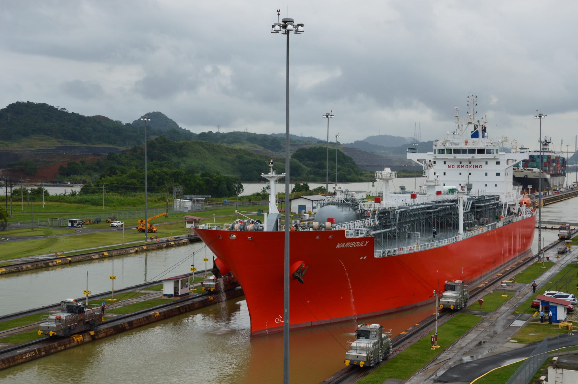 A large red tanker ship guided through a canal lock chamber by rail-mounted locomotives