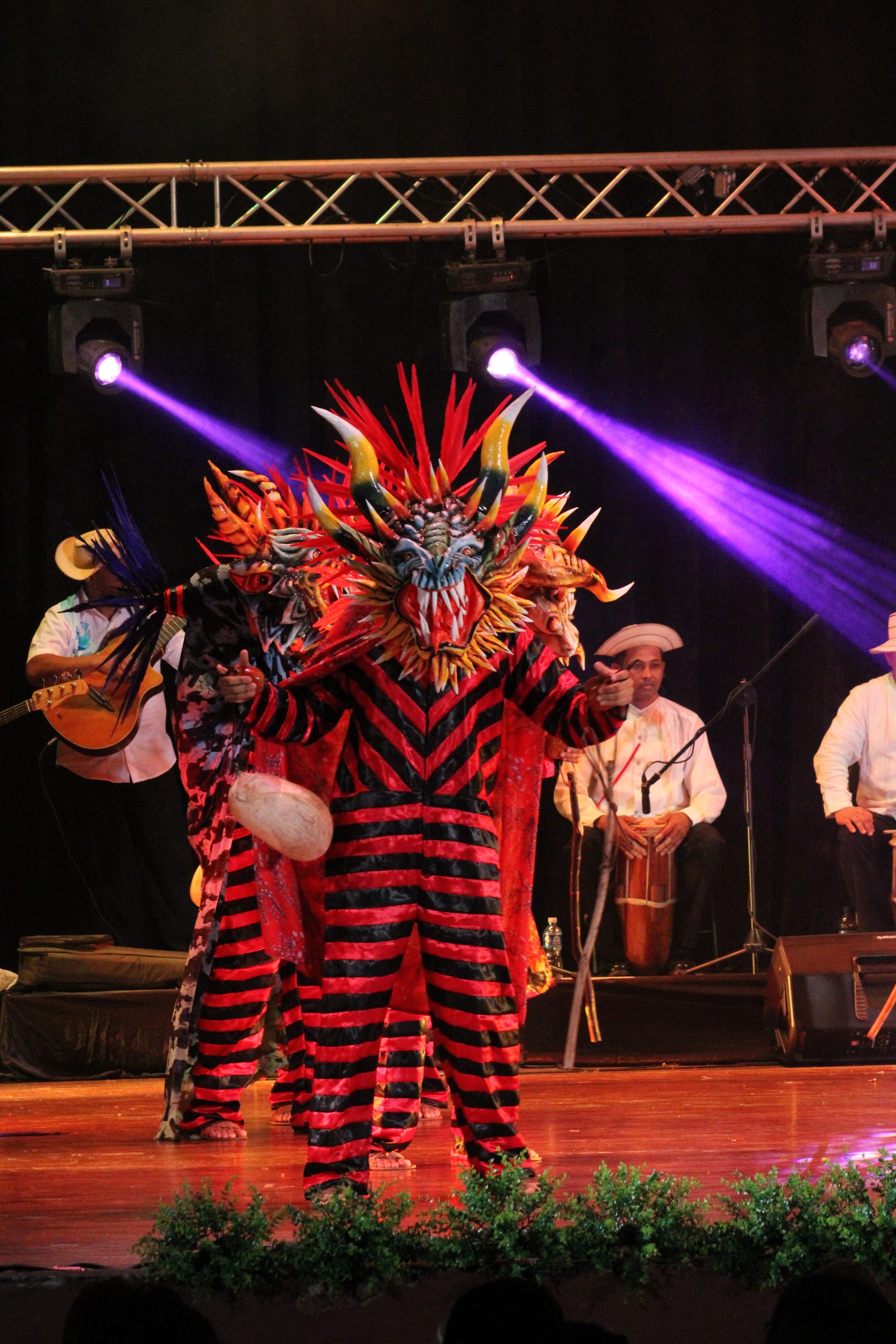A dancer in a red and black striped costume and a huge horned devil mask with red feathers and bared teeth performs on a stage, with musicians in straw hats and a drummer behind