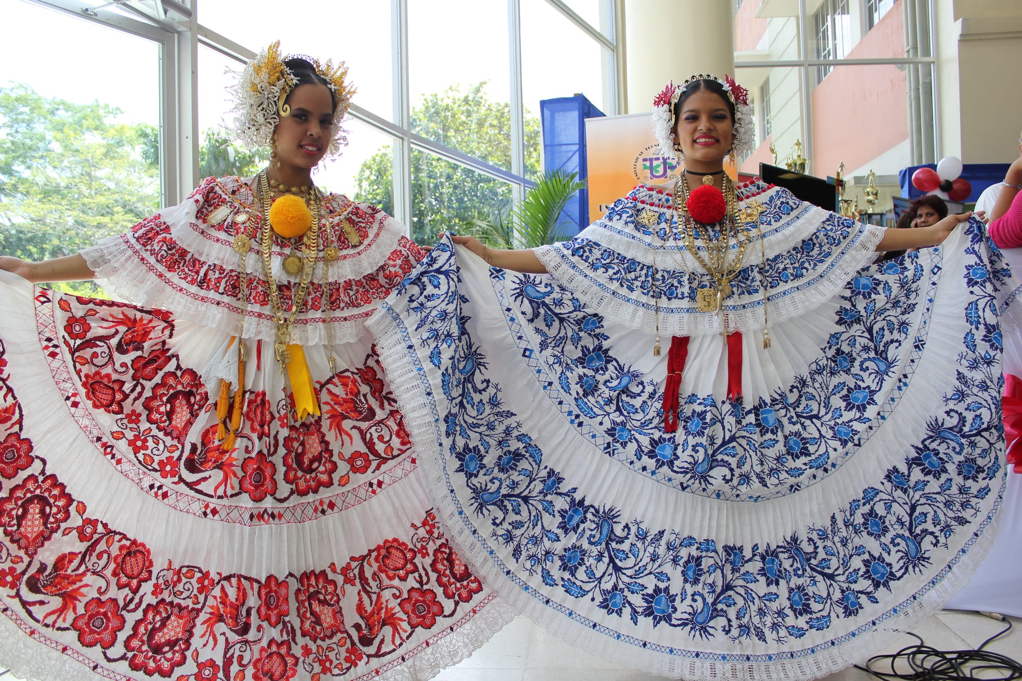 Two young women in white polleras, one embroidered in red and one in blue, spread their wide skirts, wearing gold chains, large pompoms and tall white headdresses