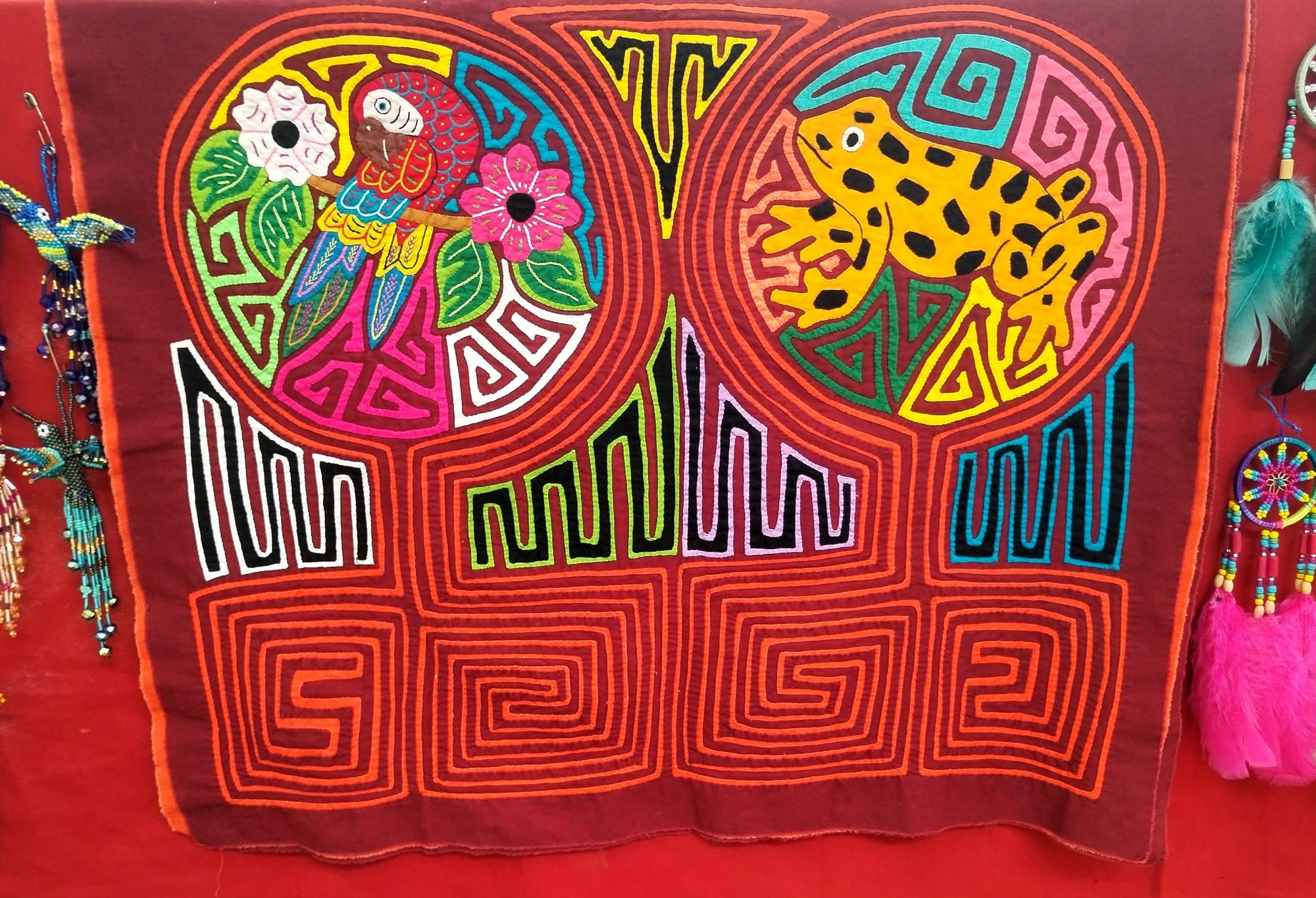 A handmade textile panel in deep red cloth with layered cut-out designs of a macaw among flowers and a spotted yellow frog, above maze-like geometric patterns in orange, green, black and blue