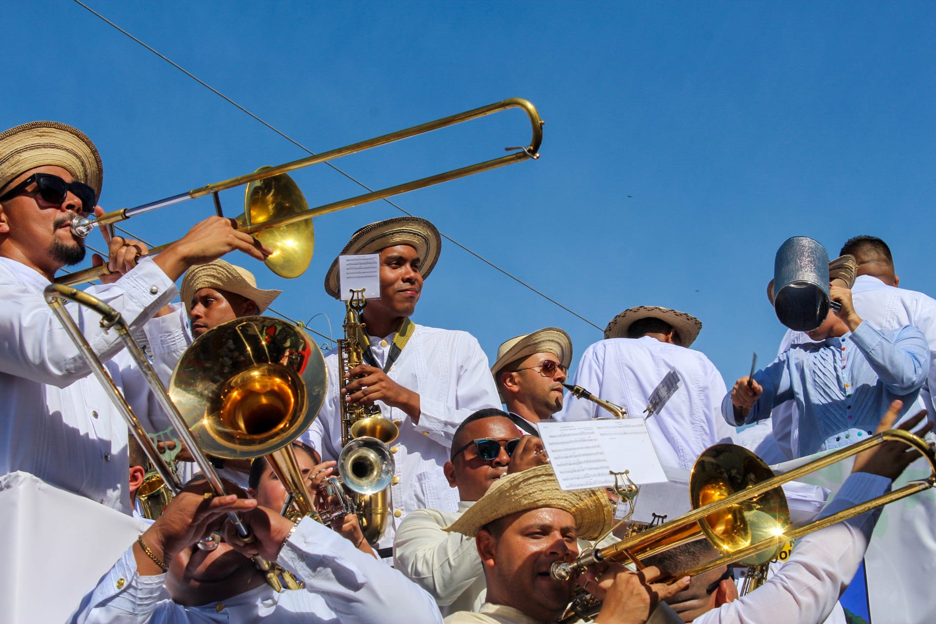 A brass band of men in white shirts and straw hats play trombones and a saxophone against a clear blue sky