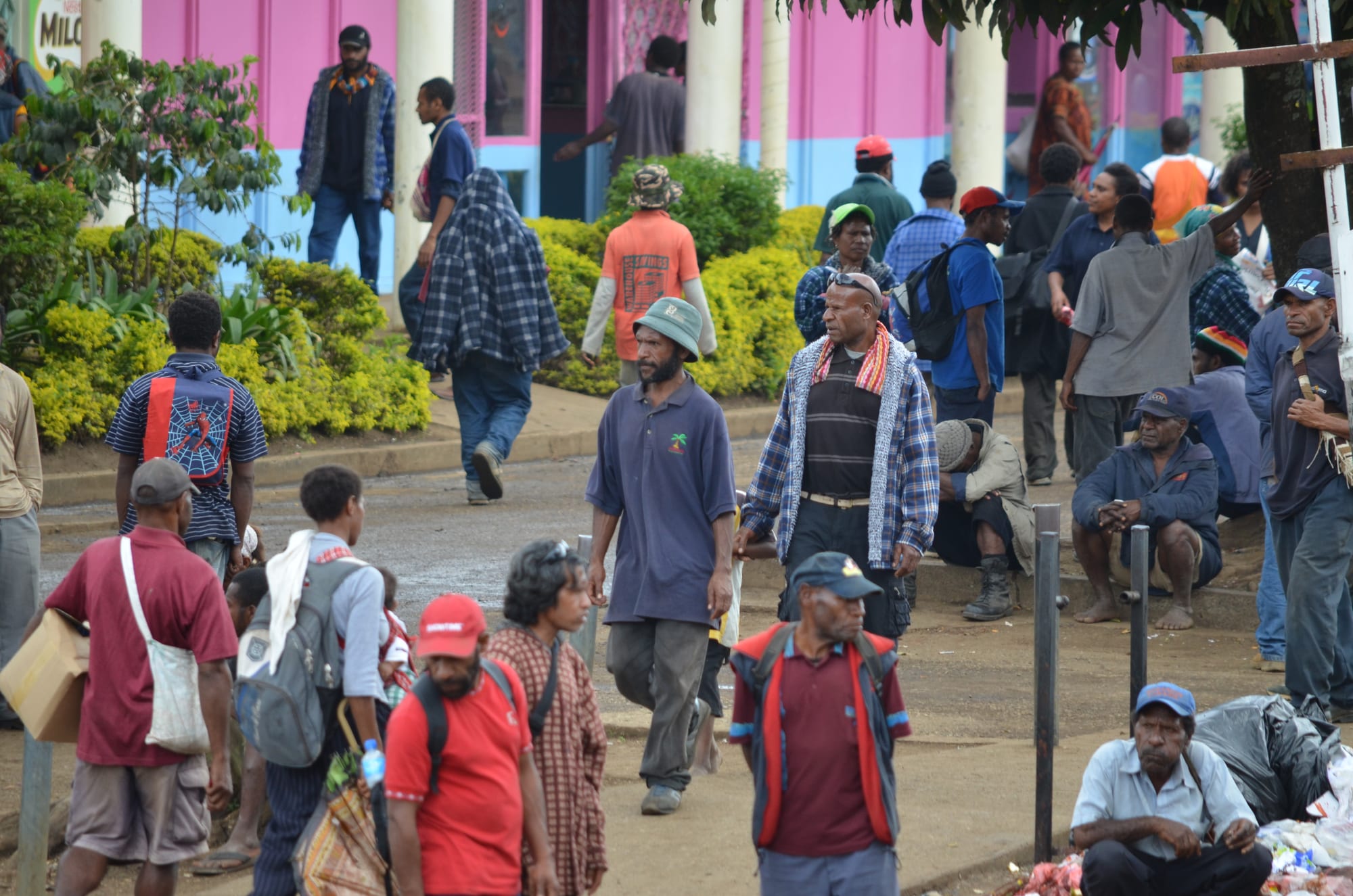 A crowded street plaza in Goroka with people walking, carrying bags and gathering near pink and blue painted buildings