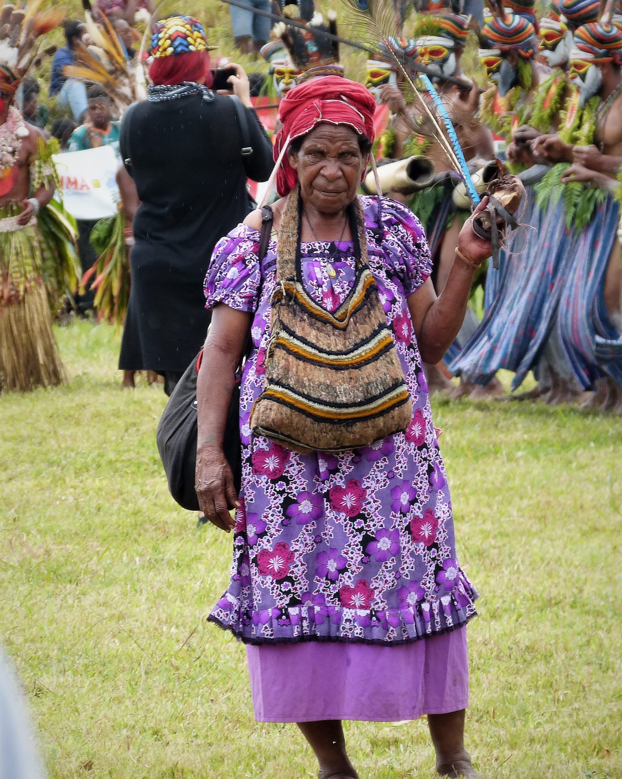 An older woman in a red headscarf and purple floral dress wears a striped brown, yellow and black string bag hung from her neck, with painted dancers and onlookers behind her on a grassy field