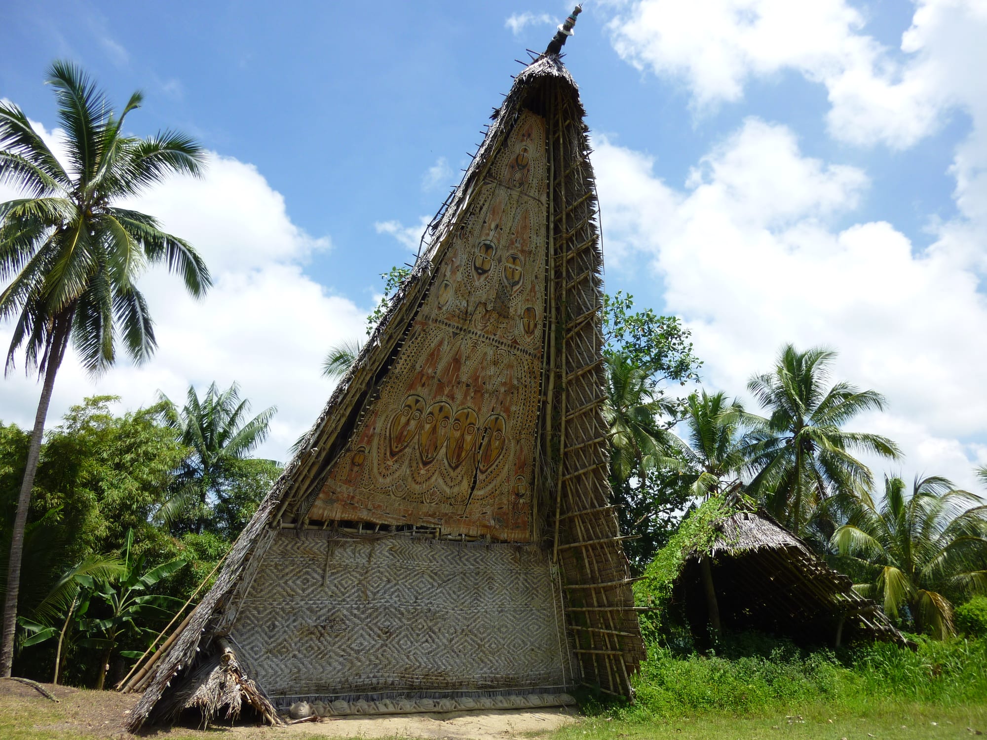 A tall thatched spirit house with a steeply pointed gable covered in painted faces and patterns, standing among coconut palms under a blue sky