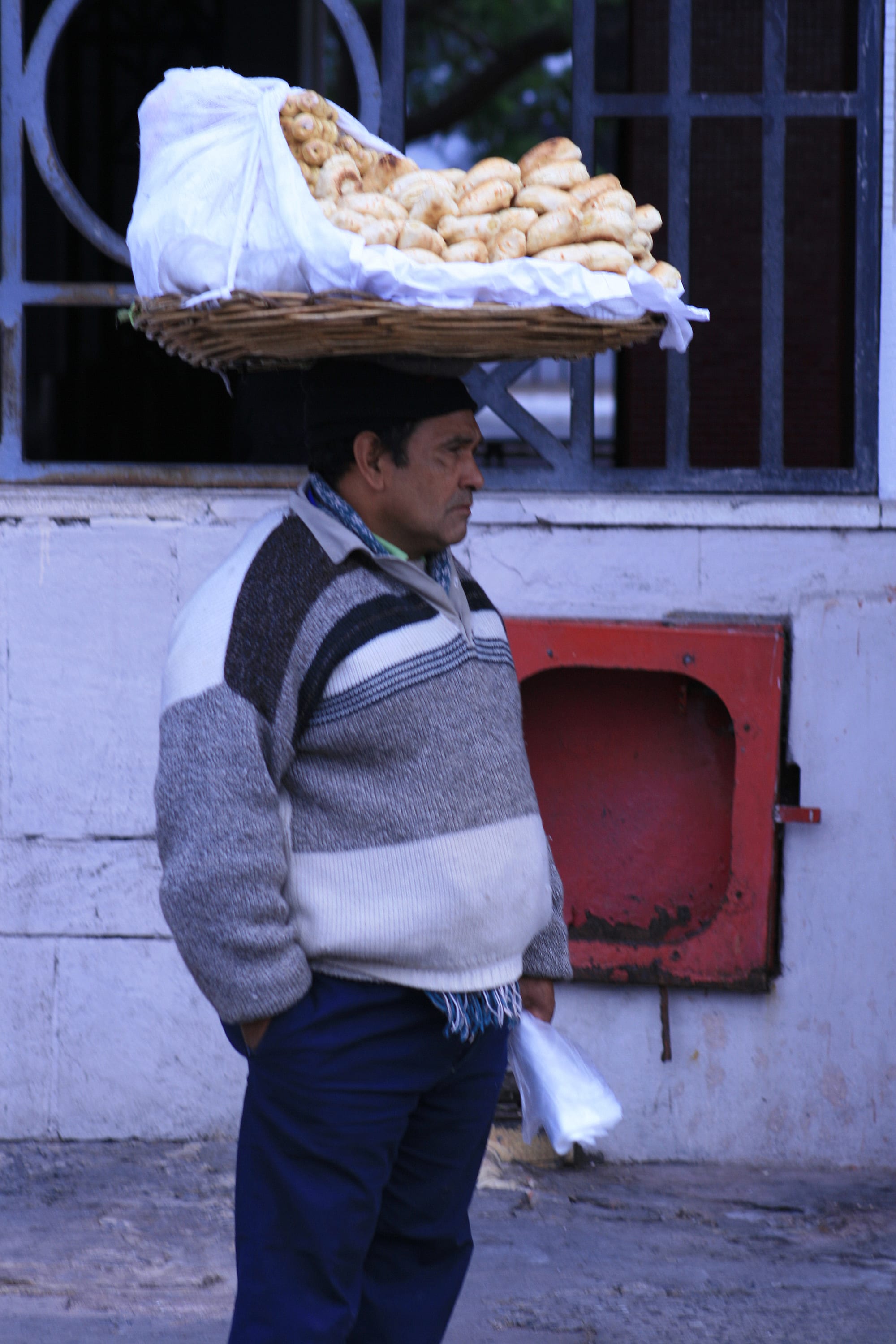 A man carrying a large woven basket of chipa bread rings on his head while walking along a street in Asunción