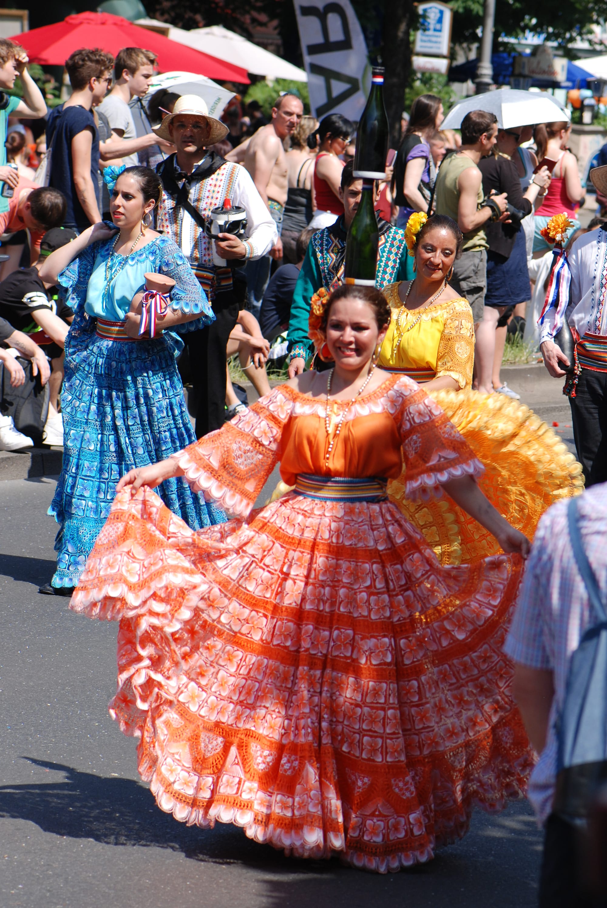Women in full, lace-patterned dresses in orange, yellow and blue dance in a street parade, one smiling with two bottles balanced on her head, as crowds watch from the pavement