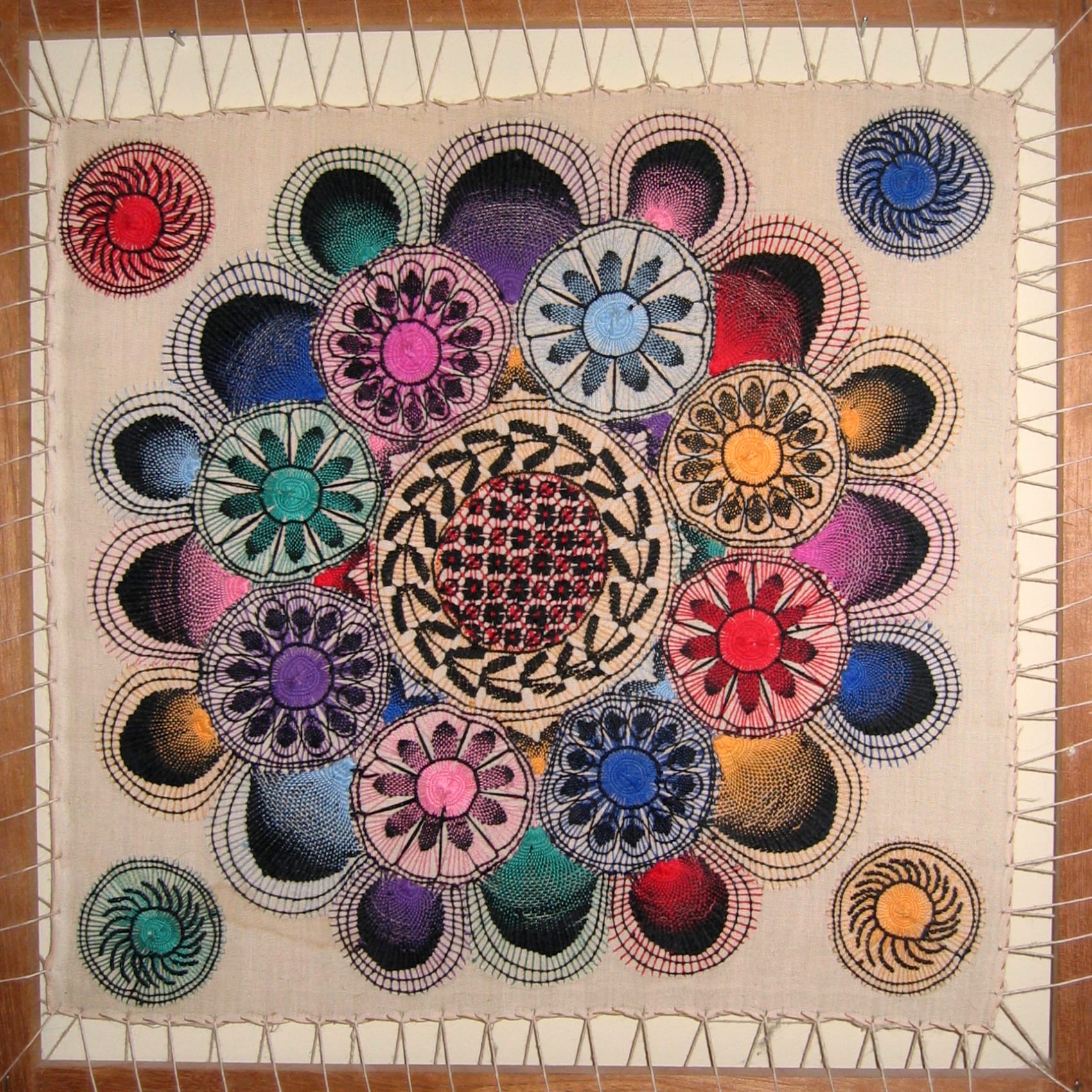 A square of cream cloth stretched on a wooden frame, covered in dozens of round lace medallions in pink, blue, green, purple, orange and black, radiating like spider webs