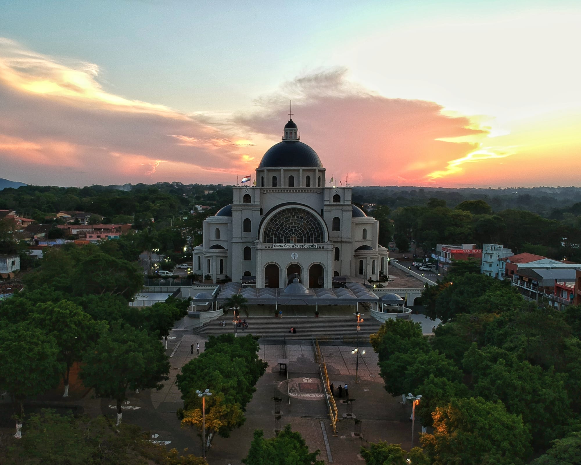 An aerial view at sunset of a large white domed basilica with a wide flight of steps and a tree-lined square in front, surrounded by low buildings and green woodland under a pink and orange sky