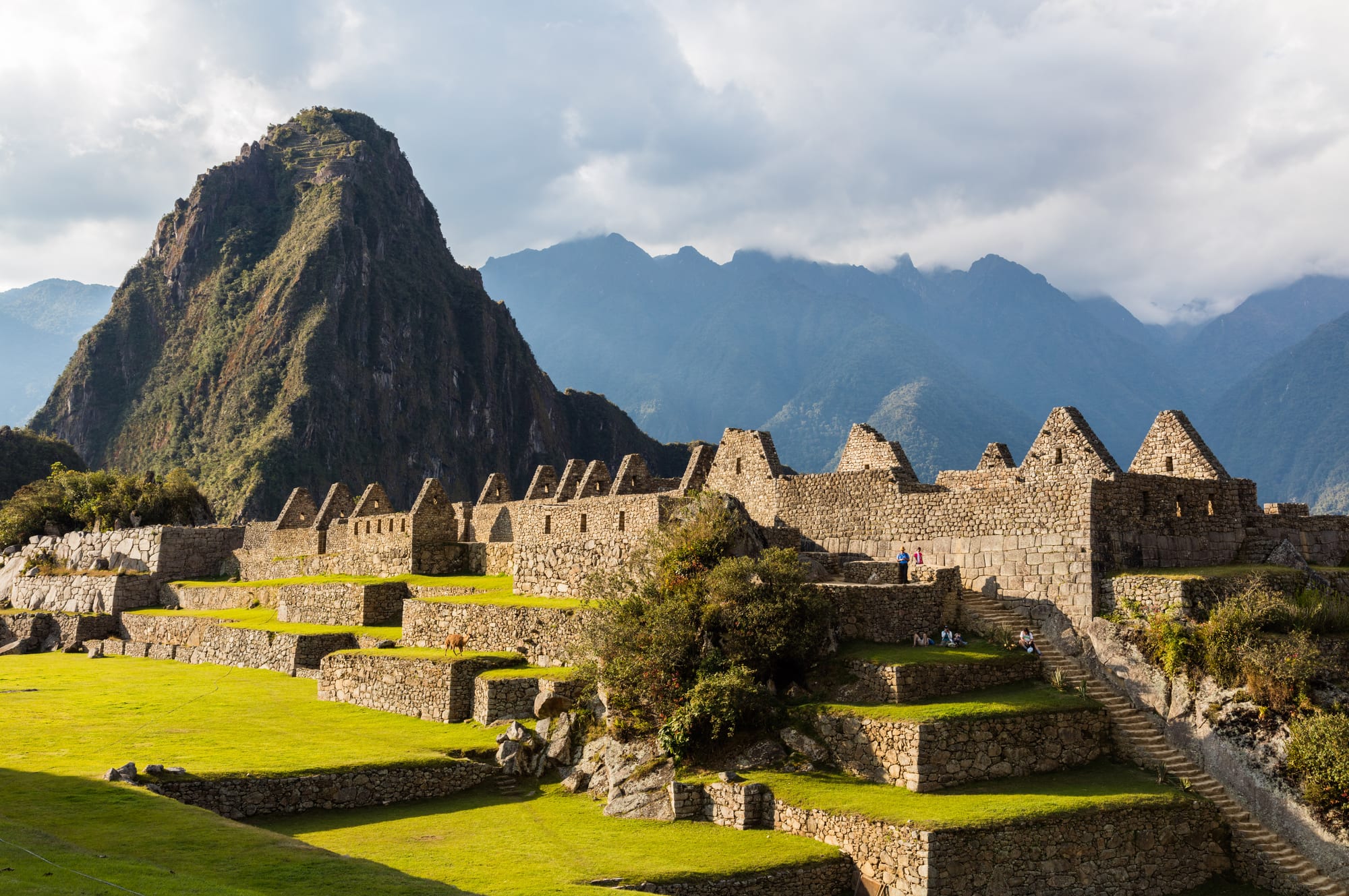 Ancient stone ruins on a green mountain terrace surrounded by steep peaks and clouds