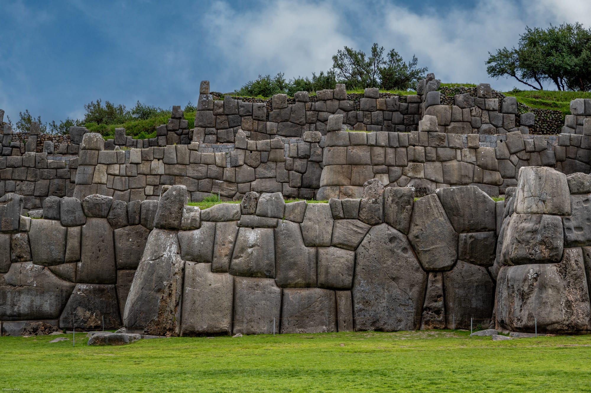 Three tiers of massive grey stone walls at Sacsayhuamán, built of huge irregular blocks fitted tightly together, above a green lawn under a cloudy sky