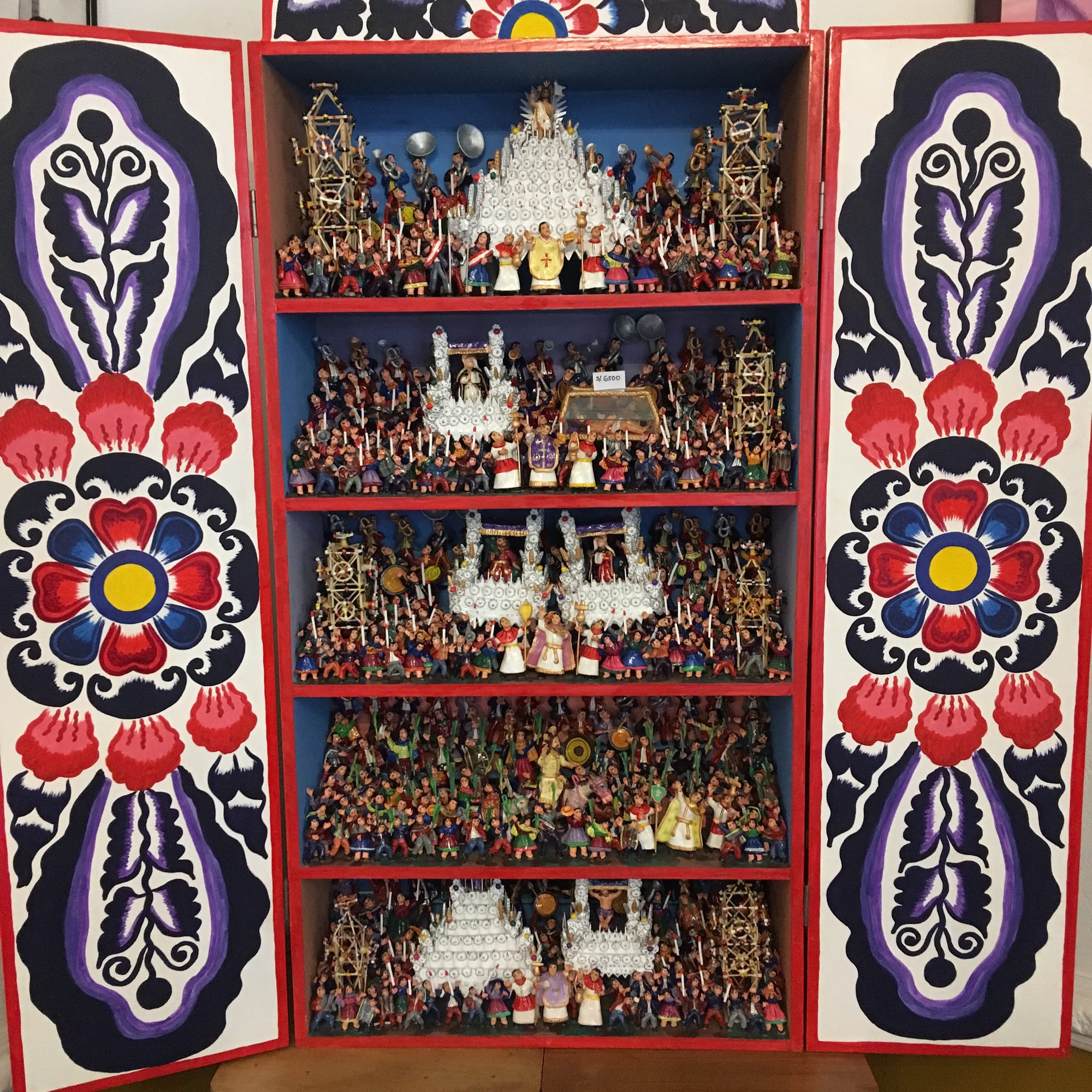 An open wooden cabinet with painted floral doors, its five shelves crowded with tiny colourful figures forming religious processions with white candle-covered platforms
