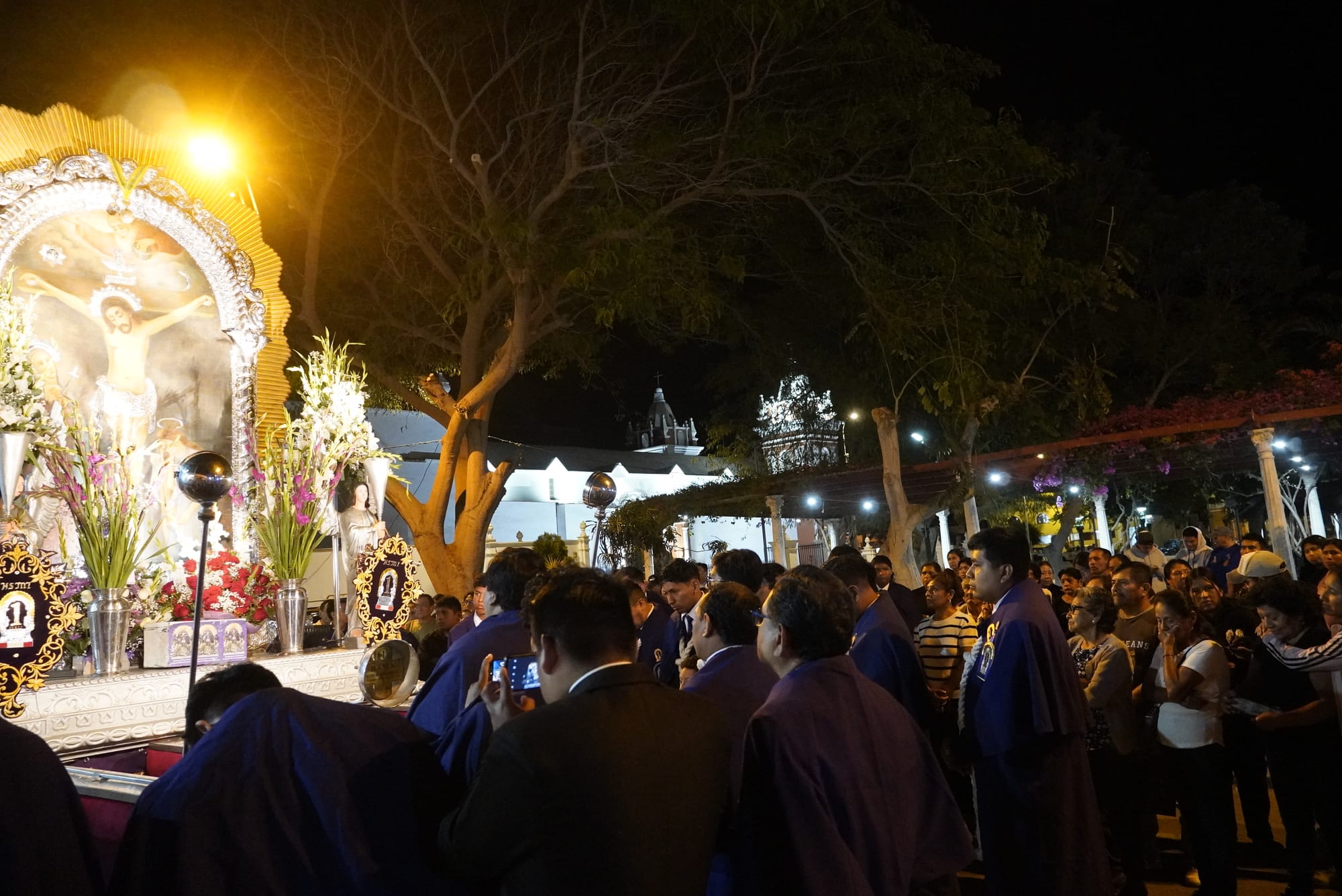 At night, a brightly lit processional platform bearing an image of the crucified Christ, framed in silver and flowers, is surrounded by men in purple habits and a large crowd of onlookers