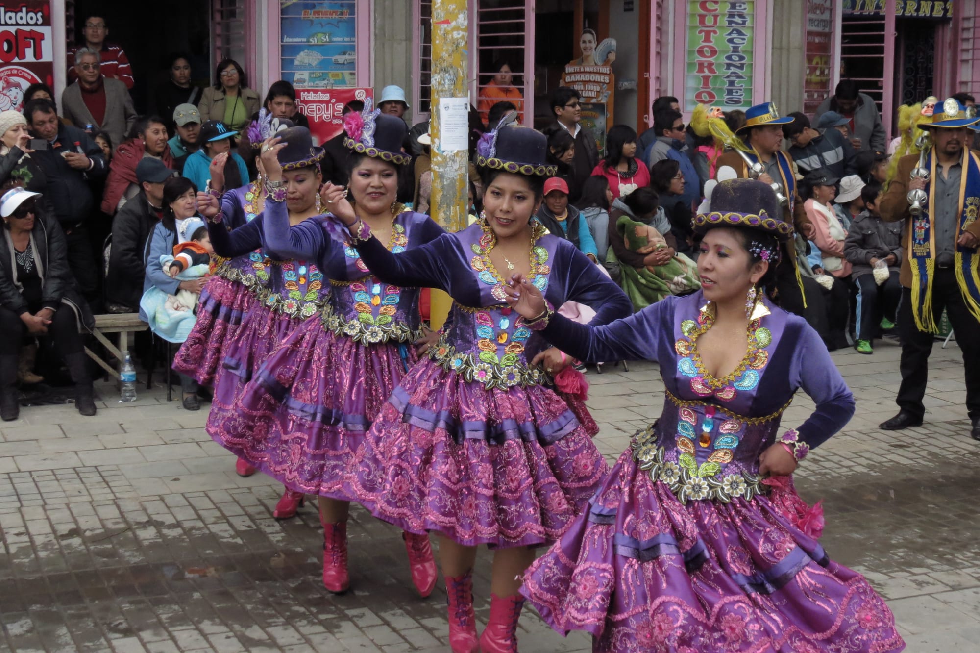 A line of women dancers in bowler hats and shimmering purple embroidered costumes with full skirts dance along a paved street as crowds watch from the pavement
