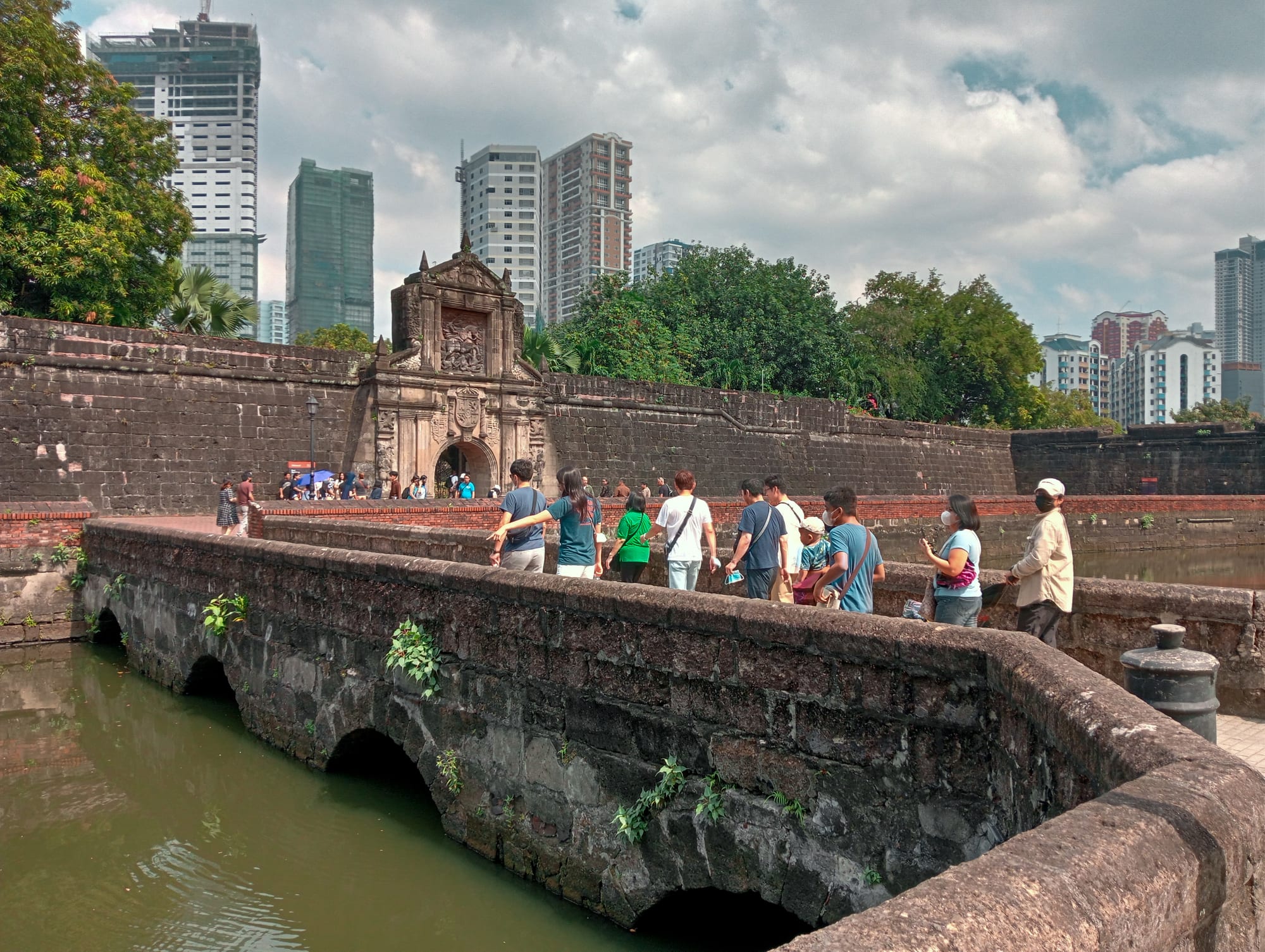 An ornate stone fort gateway across a moat bridge, modern skyscrapers behind