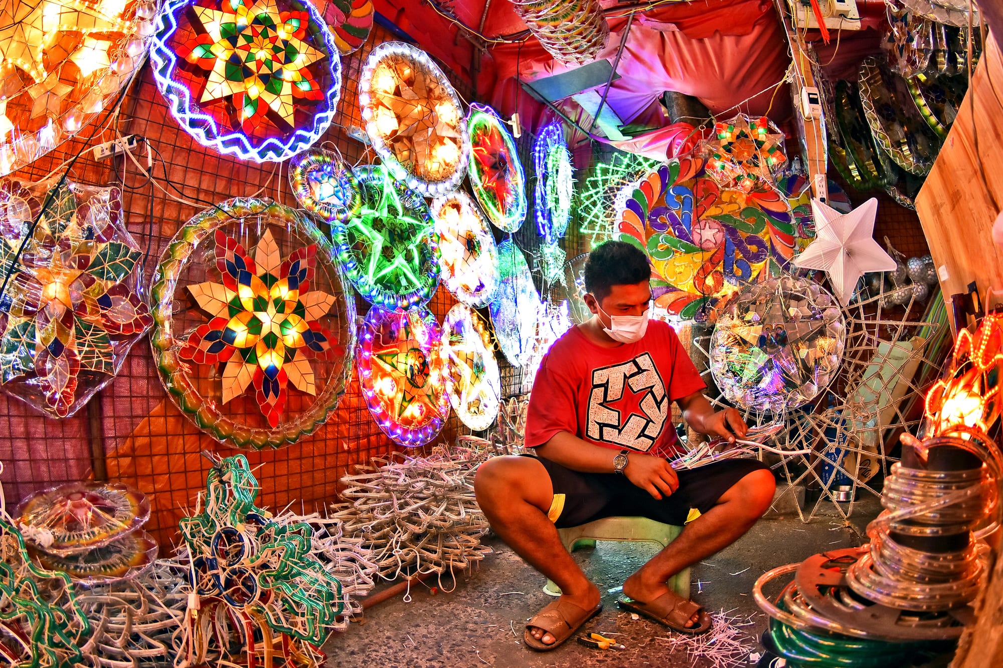 A man in a red T-shirt and face mask sits on a small stool assembling a lantern frame, surrounded by brightly lit star and circular lanterns in many colours