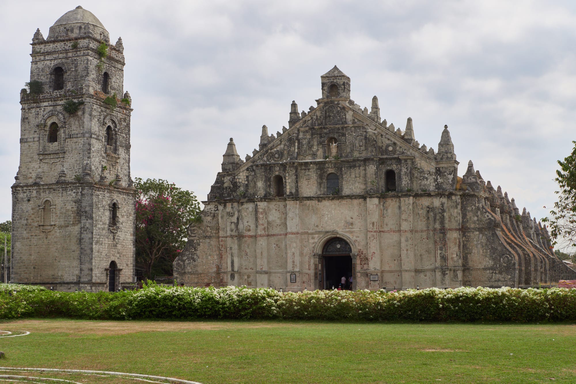 A weathered stone church with a stepped gable and massive buttresses along its side, with a separate square stone bell tower to the left and a lawn and flowering hedge in front