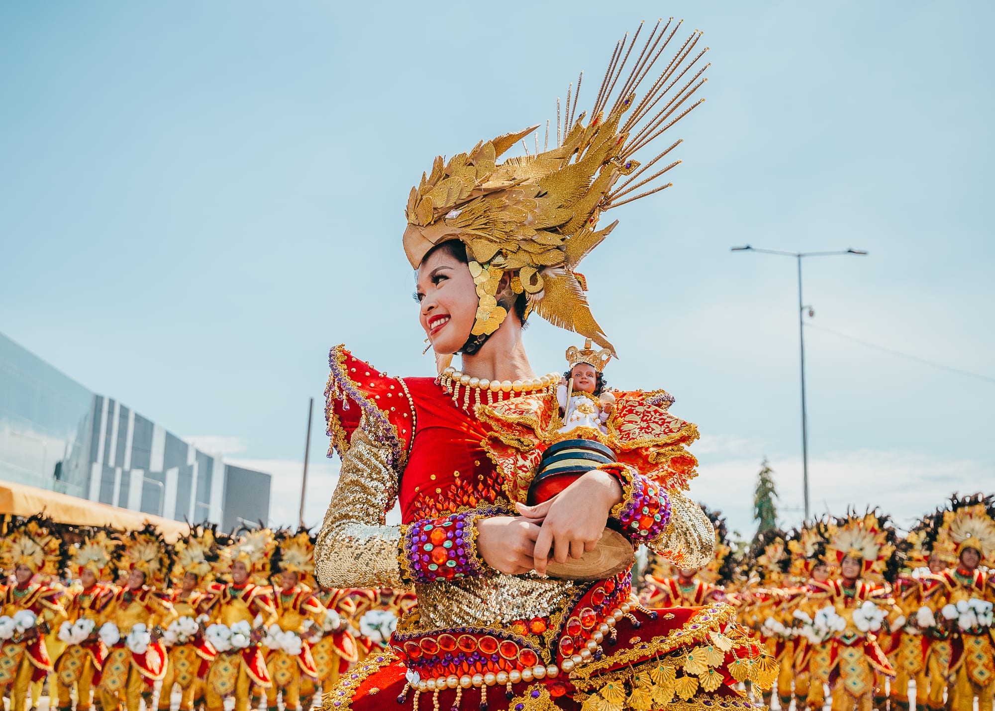A smiling dancer in a red and gold costume and a tall golden feathered headdress holds a small image of the Santo Niño, with rows of costumed dancers behind her