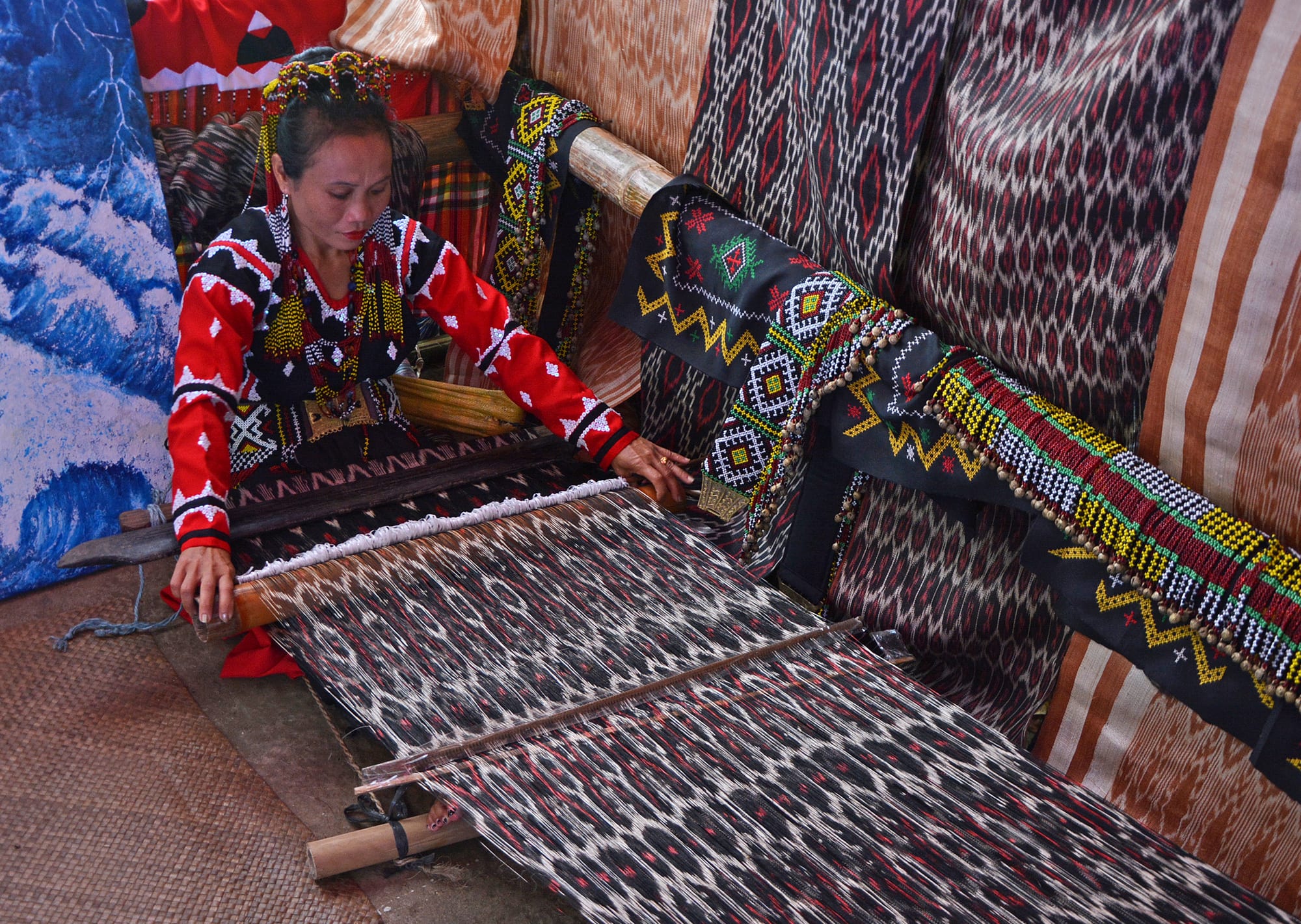 A woman in a red and black beaded blouse sits at a floor loom weaving a dark cloth patterned with pale diamond motifs, surrounded by hanging textiles and beaded belts