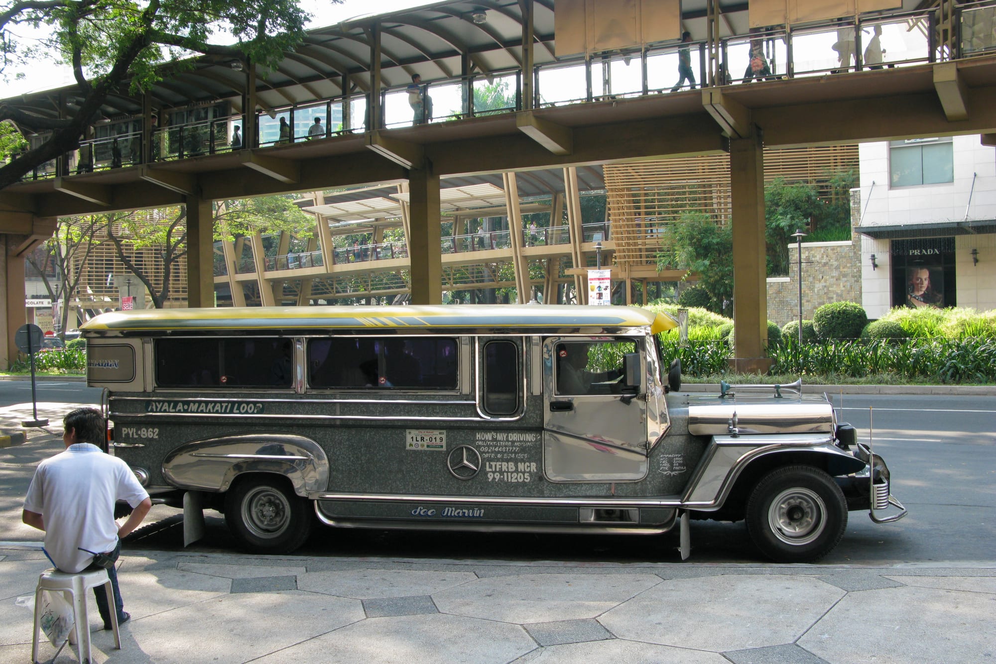 A chrome-sided jeepney with a yellow and blue roof stripe parked on a city street beneath an elevated pedestrian walkway