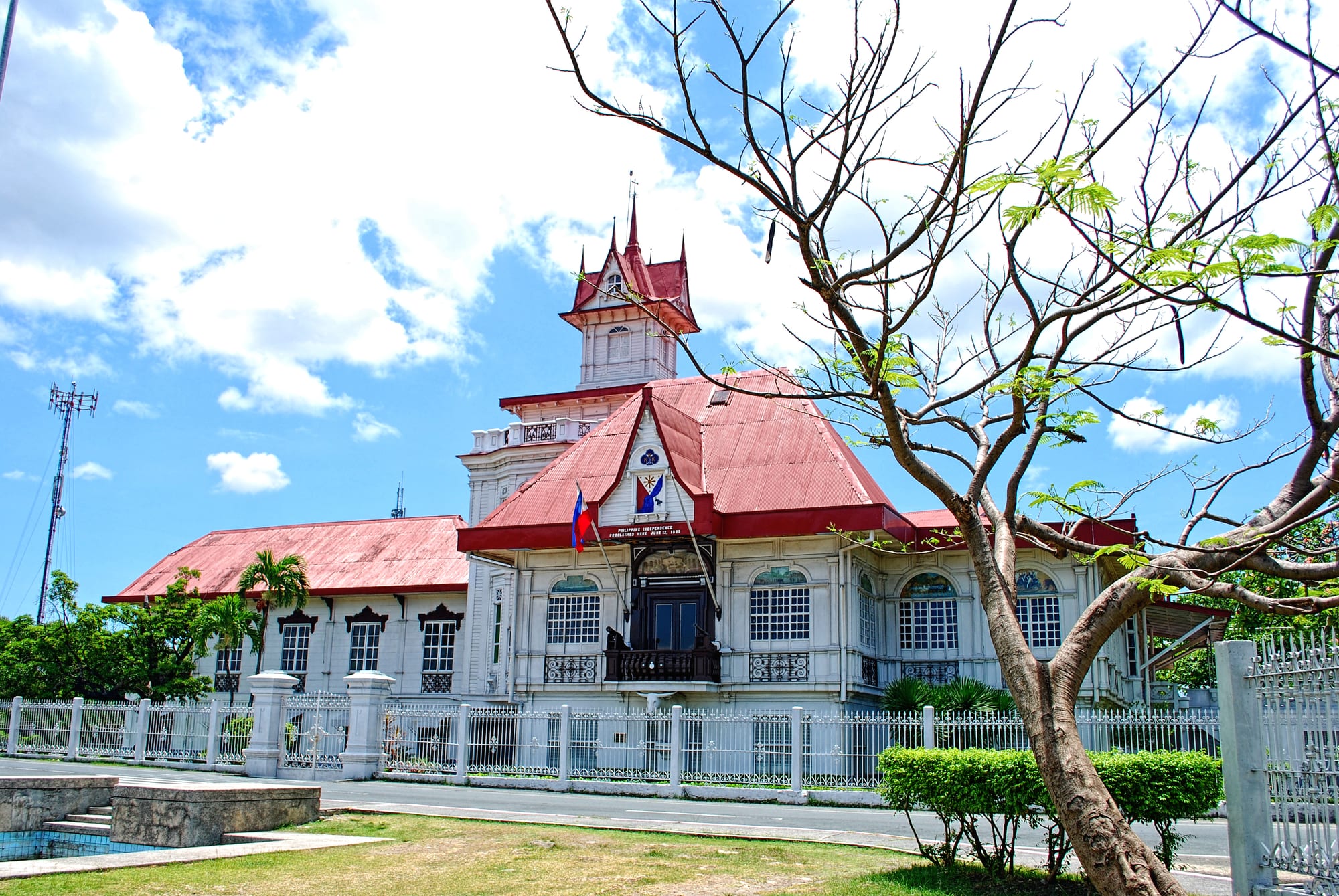 A large white mansion with red roofs and a pointed tower behind a white fence, the Philippine flag flying from its front balcony under a blue sky with clouds