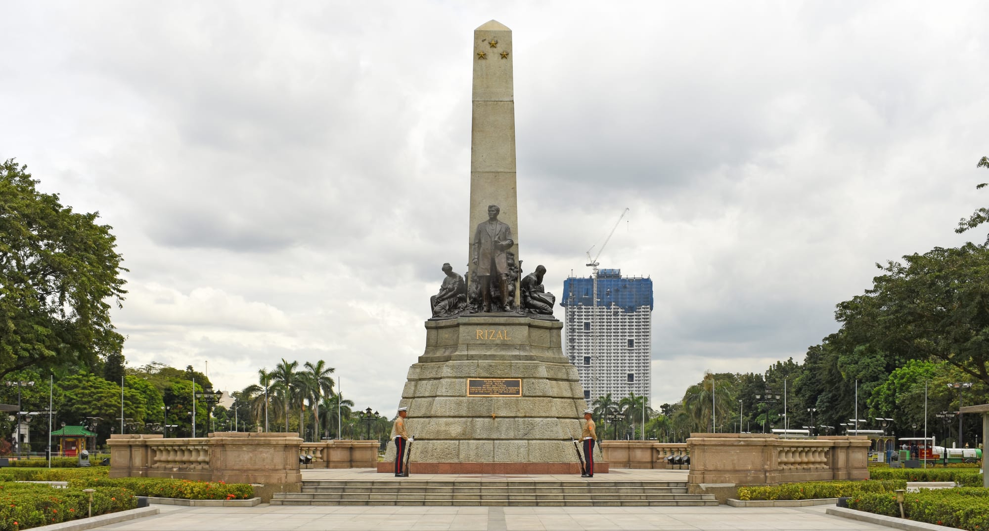 A bronze statue of José Rizal in an overcoat on a stone pedestal, guarded by two sentries, in a Manila park