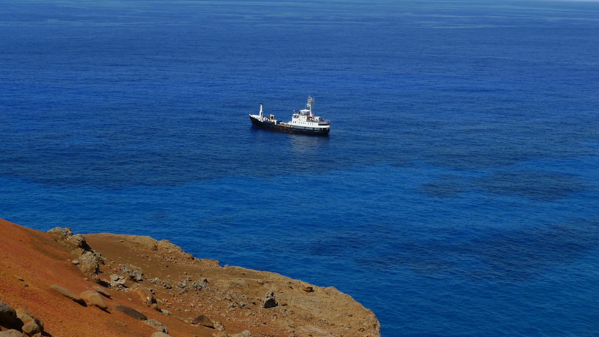 A small supply ship anchored offshore, seen from red volcanic cliffs above an empty blue ocean