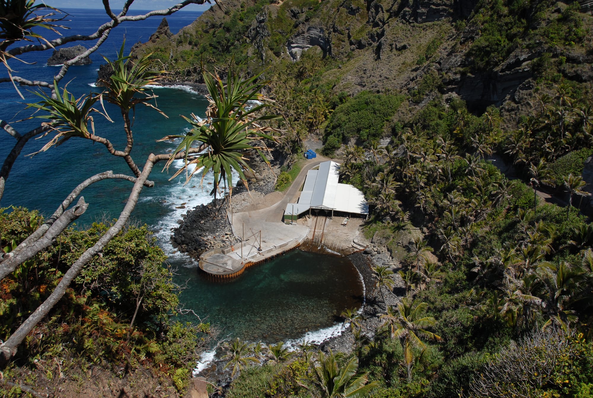 An aerial view of a small concrete boat landing and roofed shelter tucked into a rocky cove, surrounded by palms and steep cliffs