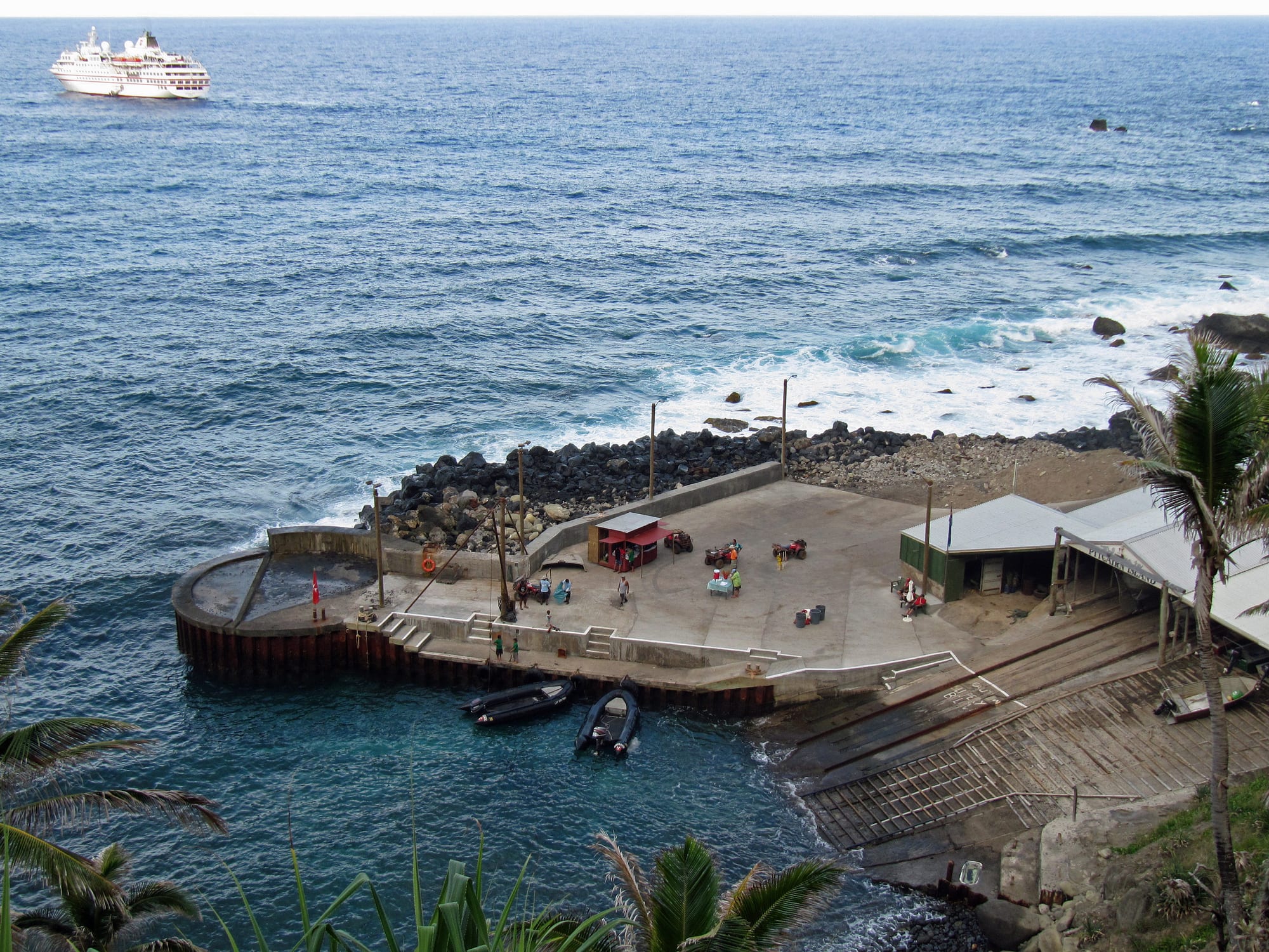 Looking down past palm fronds onto a small concrete landing jutting into the sea, with two inflatable boats tied alongside, people and quad bikes on the quay, a boat shed and slipway to the right and a white cruise ship offshore