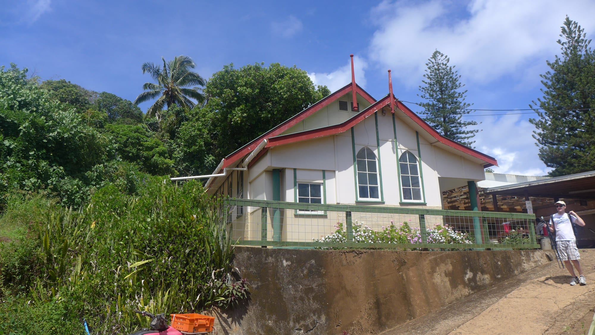 A small cream-painted building with a red-trimmed gabled roof and two tall arched windows stands behind a green railing on a raised wall, with palms and Norfolk pines behind it and a dirt road running past