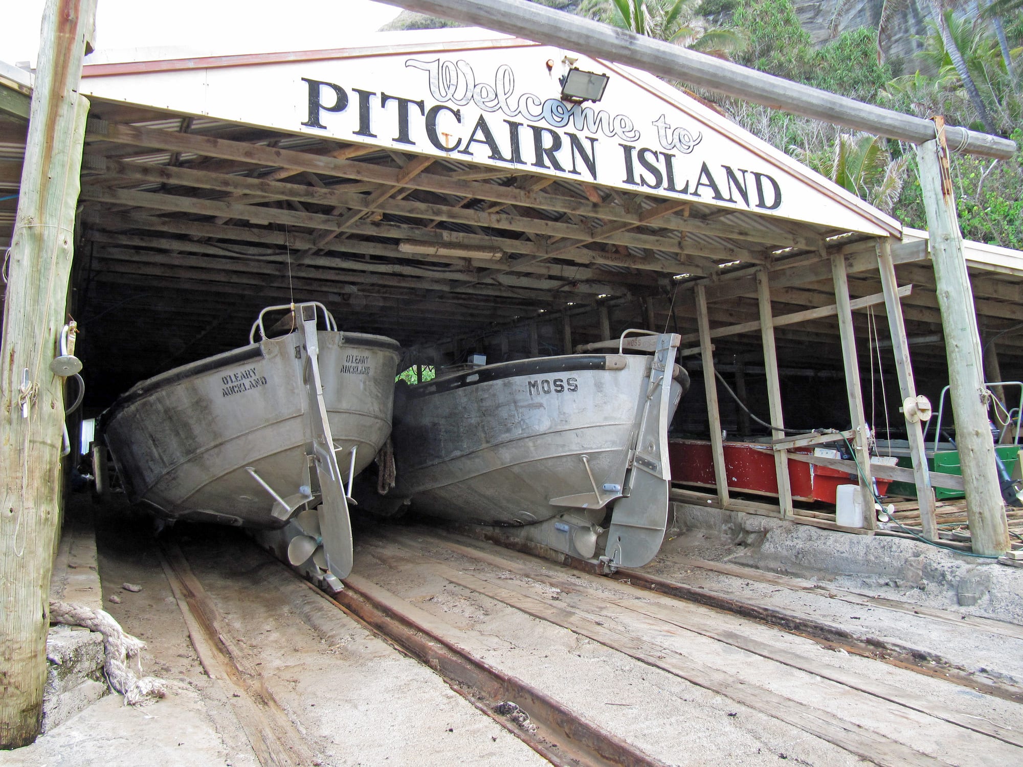 Two grey aluminium longboats rest on rails inside an open timber boat shed under a sign reading Welcome to Pitcairn Island, with steep green slopes behind