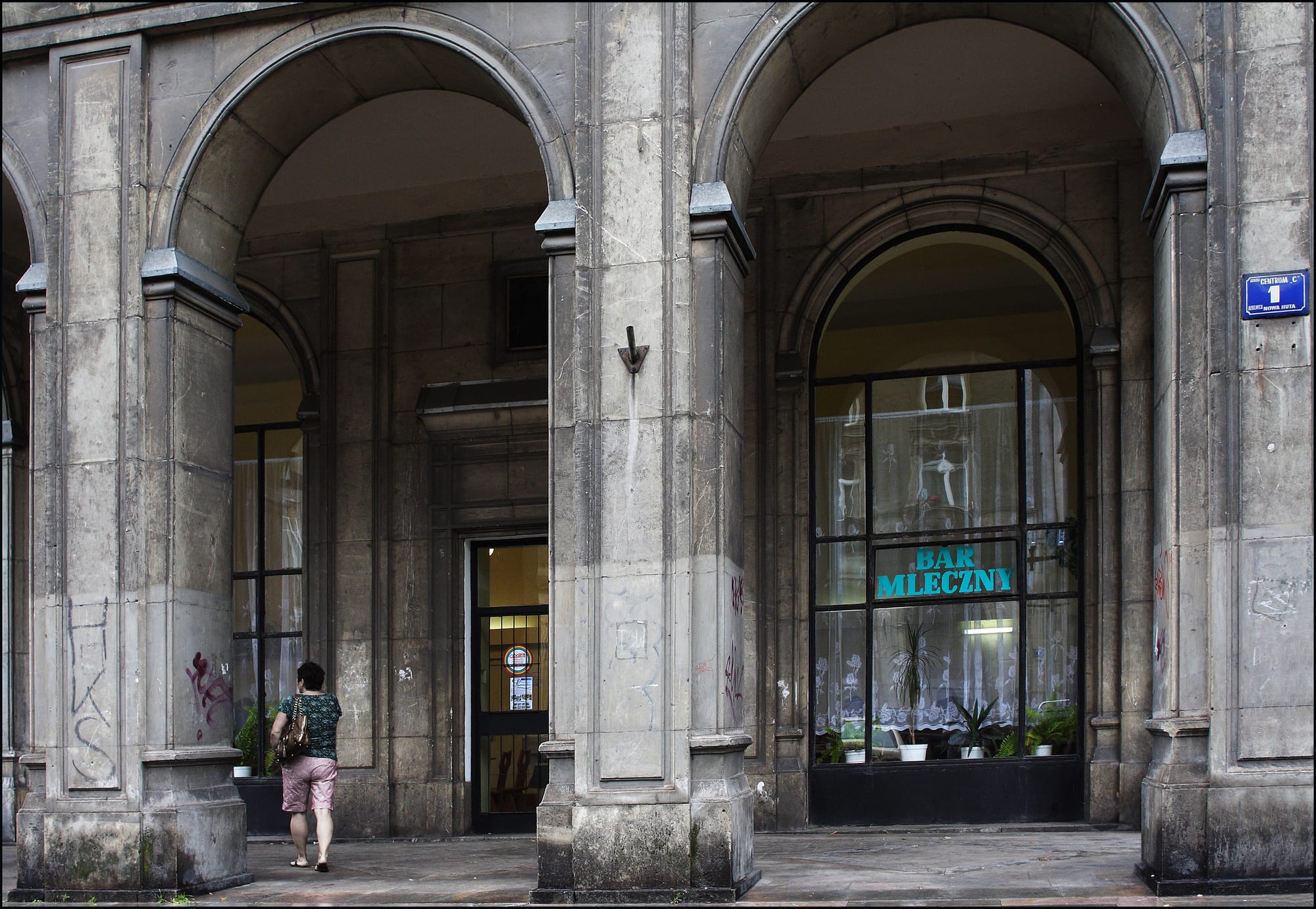 A stone archway storefront with a sign reading BAR MLECZNY, a woman walking past