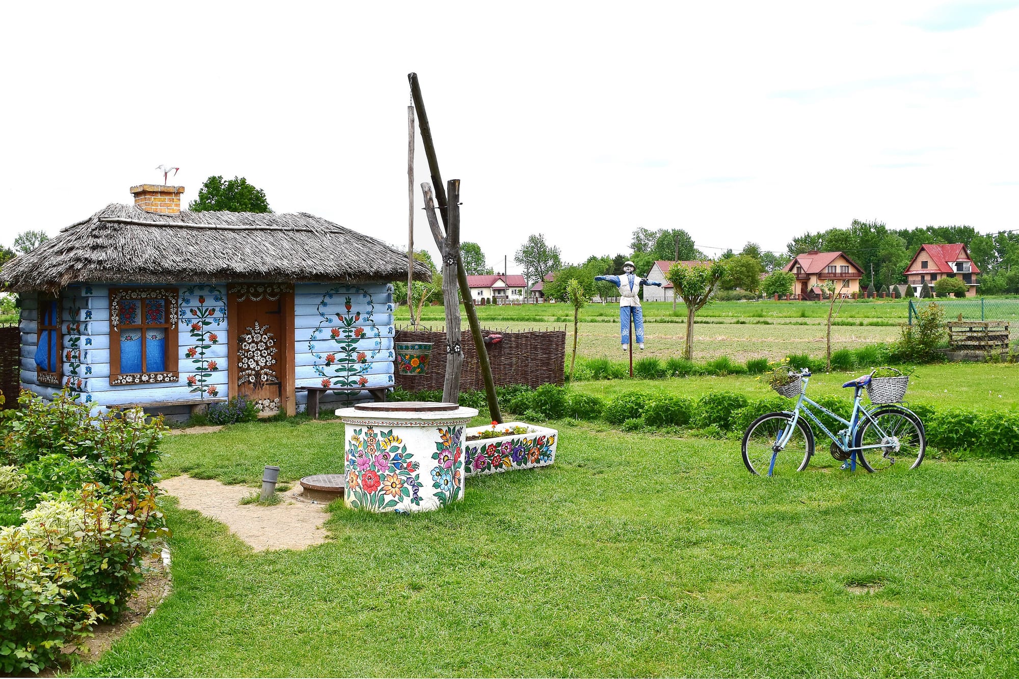 A small pale blue wooden cottage with a thatched roof, painted with red and green floral patterns around its windows and door, beside a well also painted with large flowers