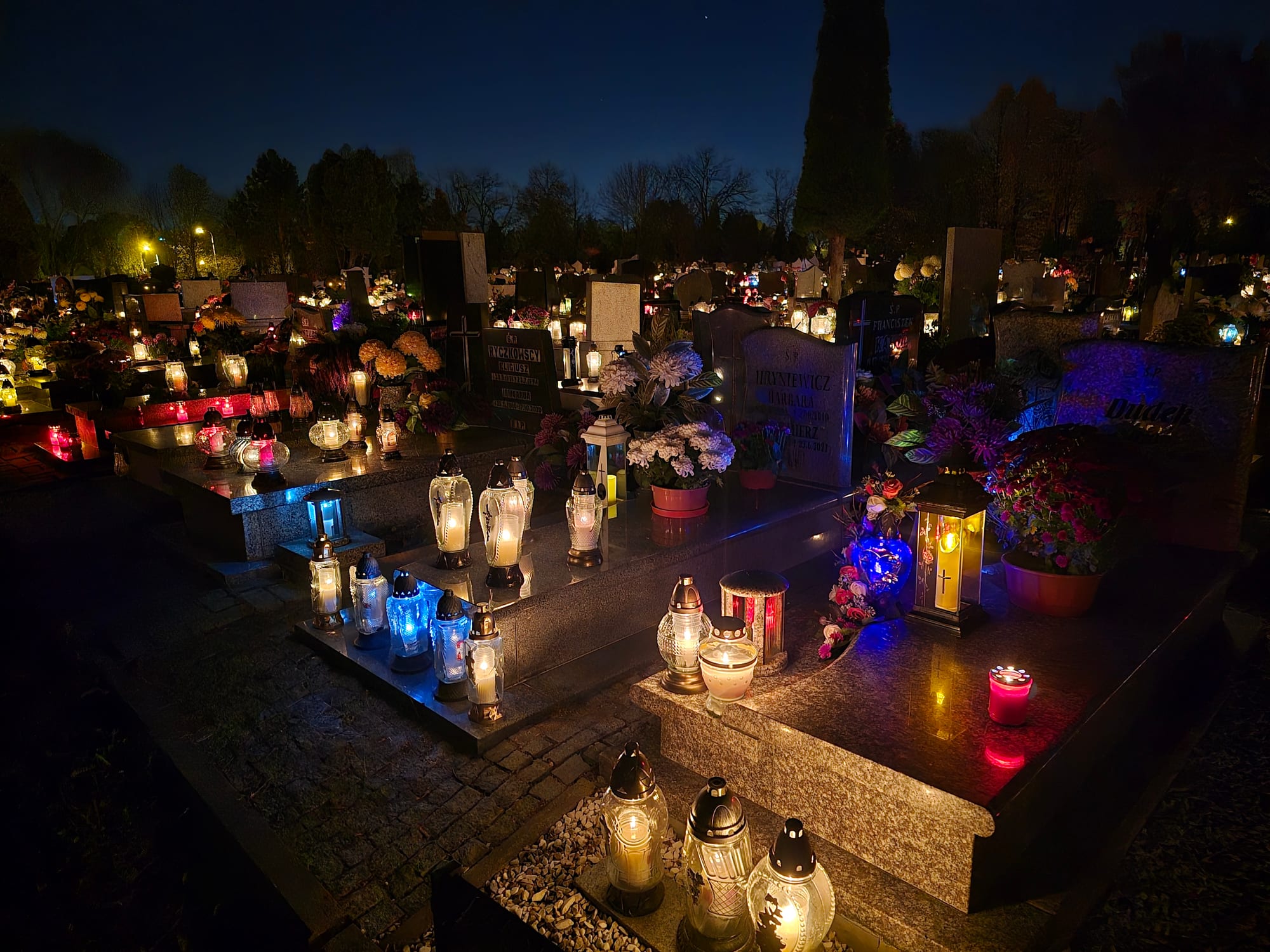 Polished stone graves at dusk covered with lit glass grave lanterns, pots of chrysanthemums and flowers, with rows of glowing candles stretching away across the cemetery