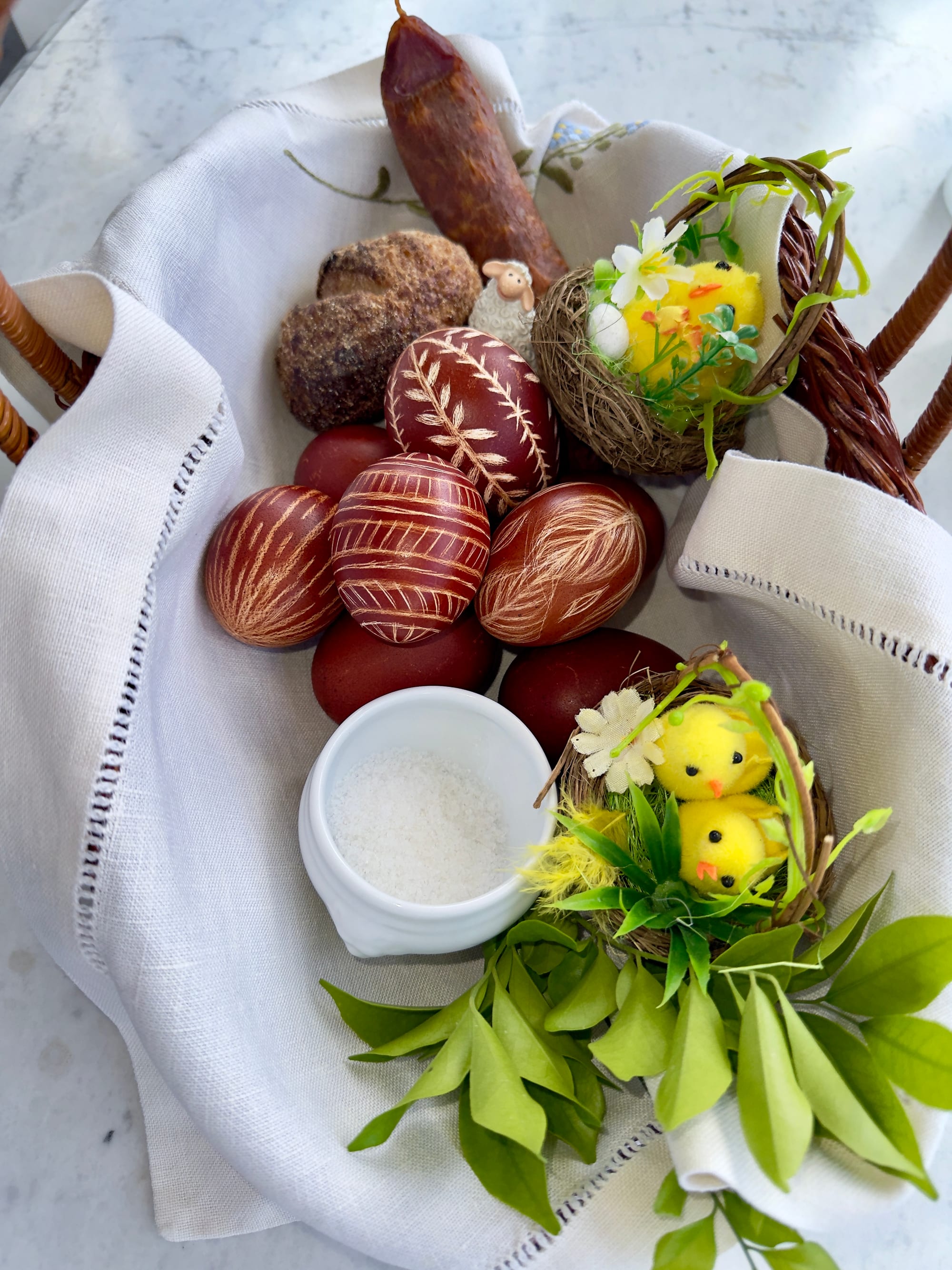 A wicker basket lined with white linen holding reddish-brown eggs etched with leaf patterns, a pot of salt, a round bread roll, a dried sausage, a small lamb figure and sprigs of greenery