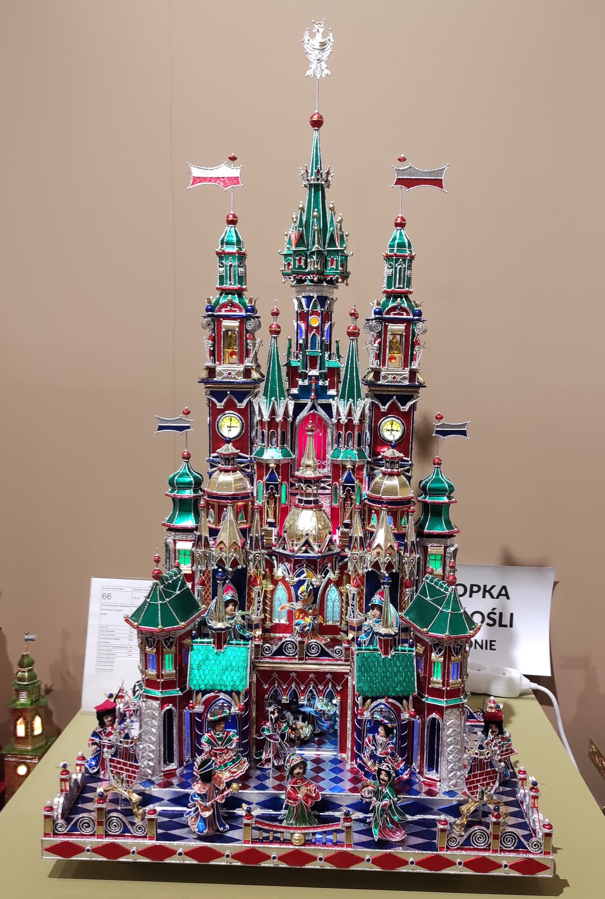 An elaborate model church with green and red spires, clock faces, tinfoil windows and Polish flags, with a nativity scene at its centre and small costumed figures in front